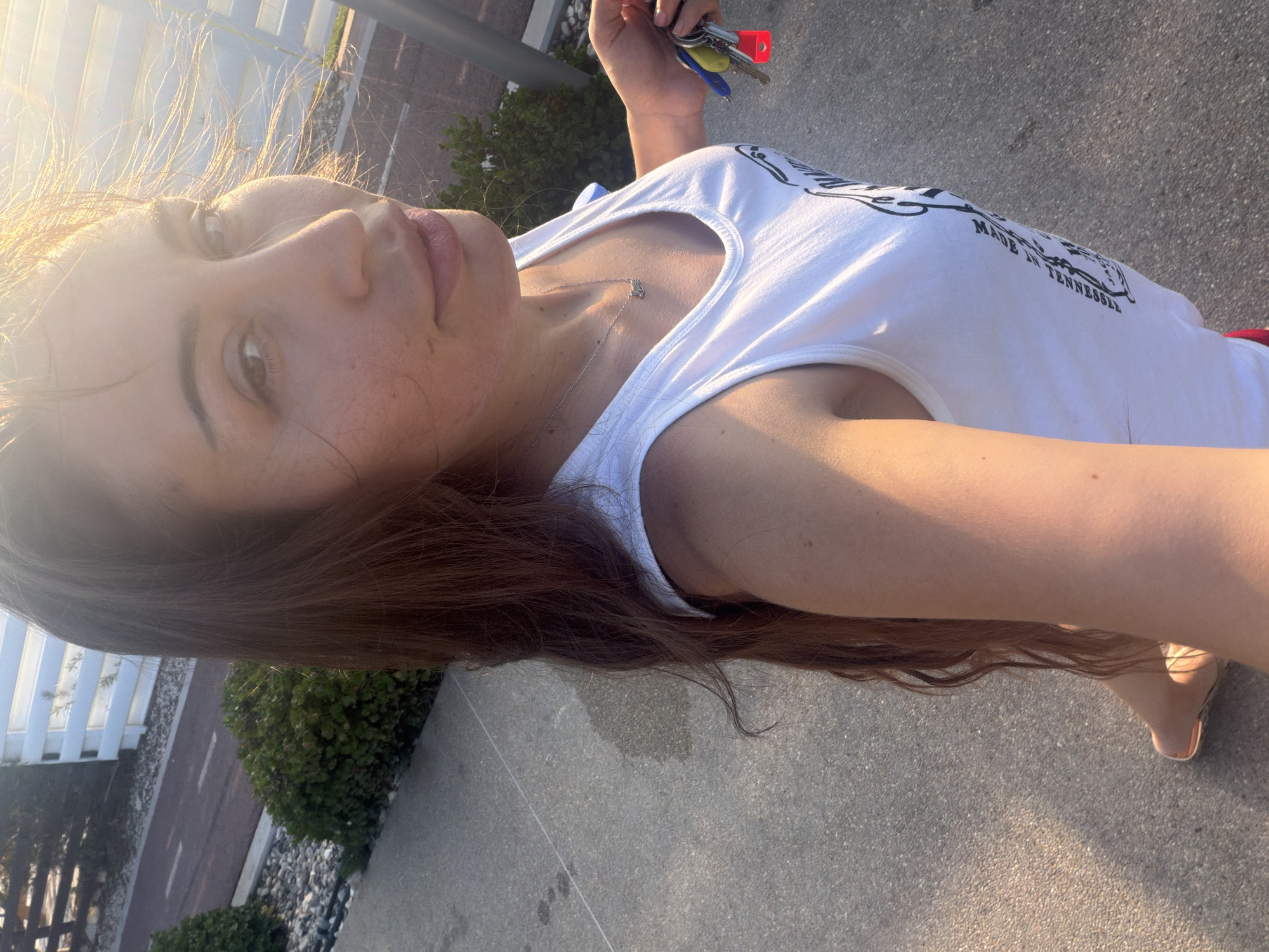 In Meneou, Cyprus, a woman with long brown hair is taking a selfie outdoors on a paved walkway. She is wearing a white tank top with a graphic print and a silver necklace. In her right hand, she holds a set of keys with a red fob. Sunlight is casting a warm glow on her face and the environment, with some wind rustling her hair. To her left, there are green bushes and a white fence bordering a path.