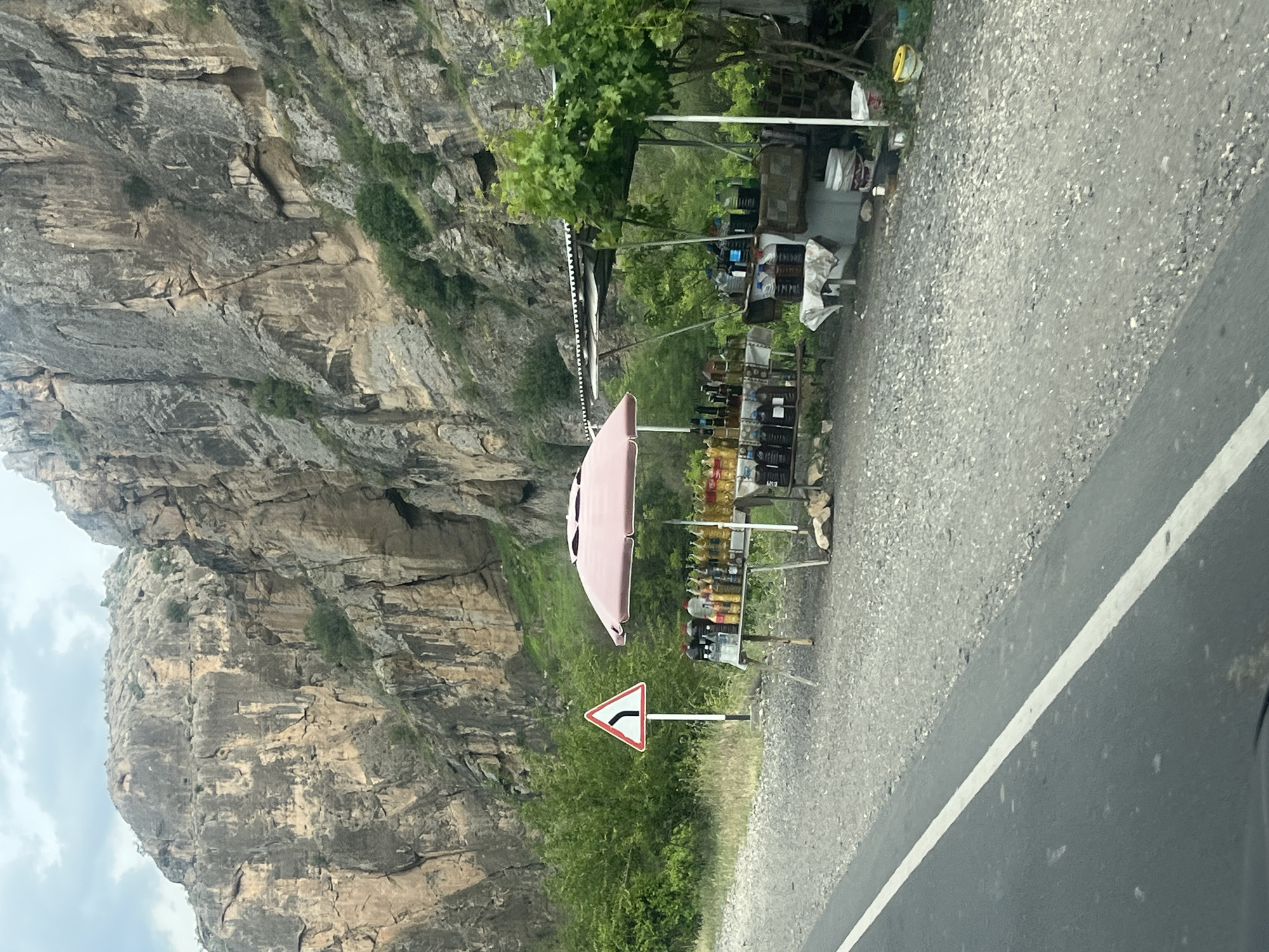 The image, oriented correctly, displays a roadside scene in a mountainous area. On the left, a towering, rugged mountain face dominates, characterized by layers of grey and brown rock formations and scattered green vegetation. The sky above the mountain is partly cloudy.
In the foreground, on the right side of the image, is an asphalt road with a solid white line marking its edge. Adjacent to the road is a gravel shoulder where a makeshift roadside vendor stall is set up. The stall is partially shaded by a light pink or beige umbrella. Numerous bottles, filled with various liquids (likely beverages, oils, or local products), are neatly arranged on shelves and tables for sale. Many bottles are transparent, showcasing contents ranging from golden to dark amber. Some items are also seen in plastic crates. Behind the stall, lush green trees and undergrowth add to the natural setting.
A white triangular warning sign with a red border and a black curved arrow, indicating a sharp left turn ahead, is clearly visible on a pole beside the road and near the vendor stall. The scene suggests a typical roadside market catering to travelers on a mountain pass. No people are visible in the frame. The lighting indicates it is daytime, possibly overcast.