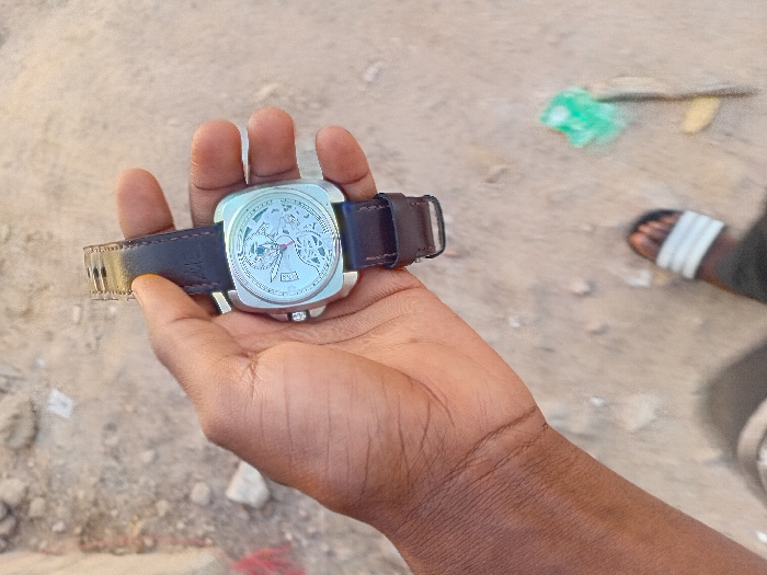 A dark-skinned right hand holds a silver-cased, square-shaped wristwatch with a skeletonized dial and a dark brown leather strap. The watch features visible internal mechanisms, a small detail at the 3 o'clock position, and a buckle on the strap. Below, a portion of a left foot wearing a black sandal with white stripes is visible on a light brown, unpaved ground. The ground surface consists of dirt and small stones, with scattered minor debris including a green plastic object and reddish fabric. The scene is outdoors in Dorayi, Nigeria.