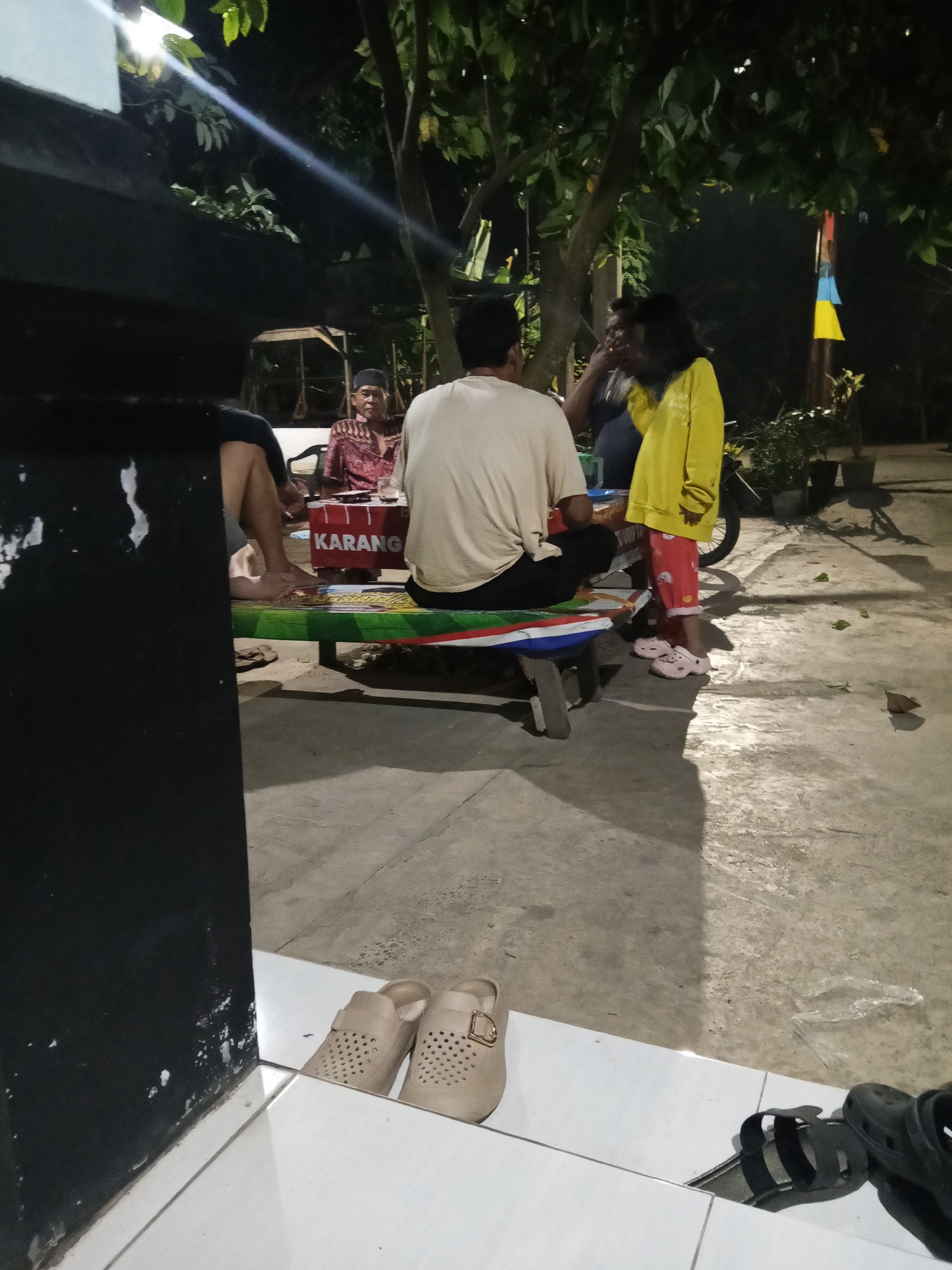 In the city of Lubuklinggau, Indonesia, a group of people is gathered outdoors at night, possibly in a residential area or a small roadside stall. The scene is dimly lit by artificial light, casting long shadows.
In the foreground, a pair of beige perforated clogs rests on a white tiled surface. To the right, a pair of black sandals is also visible on the same tiled area.
The main focus of the image is a group of four individuals seated and standing around a table. A man wearing a patterned shirt and a dark cap is seated at the table. To his right, a person in a light-colored t-shirt and dark pants is seated. A woman in a bright yellow long-sleeved shirt and pink pants with white patterns is standing to the right, facing away from the camera. Another person, whose face is obscured, is seated on the left side of the table.
The table itself is low and has a colorful, patterned cloth covering it, with the word "KARANG" prominently displayed in red lettering on a white banner at the front. The table appears to be on a wheeled platform or a cart.
The environment suggests an outdoor setting with trees and foliage in the background. There are potted plants and a bicycle wheel visible. The ground is paved with concrete. The overall atmosphere is casual and communal.