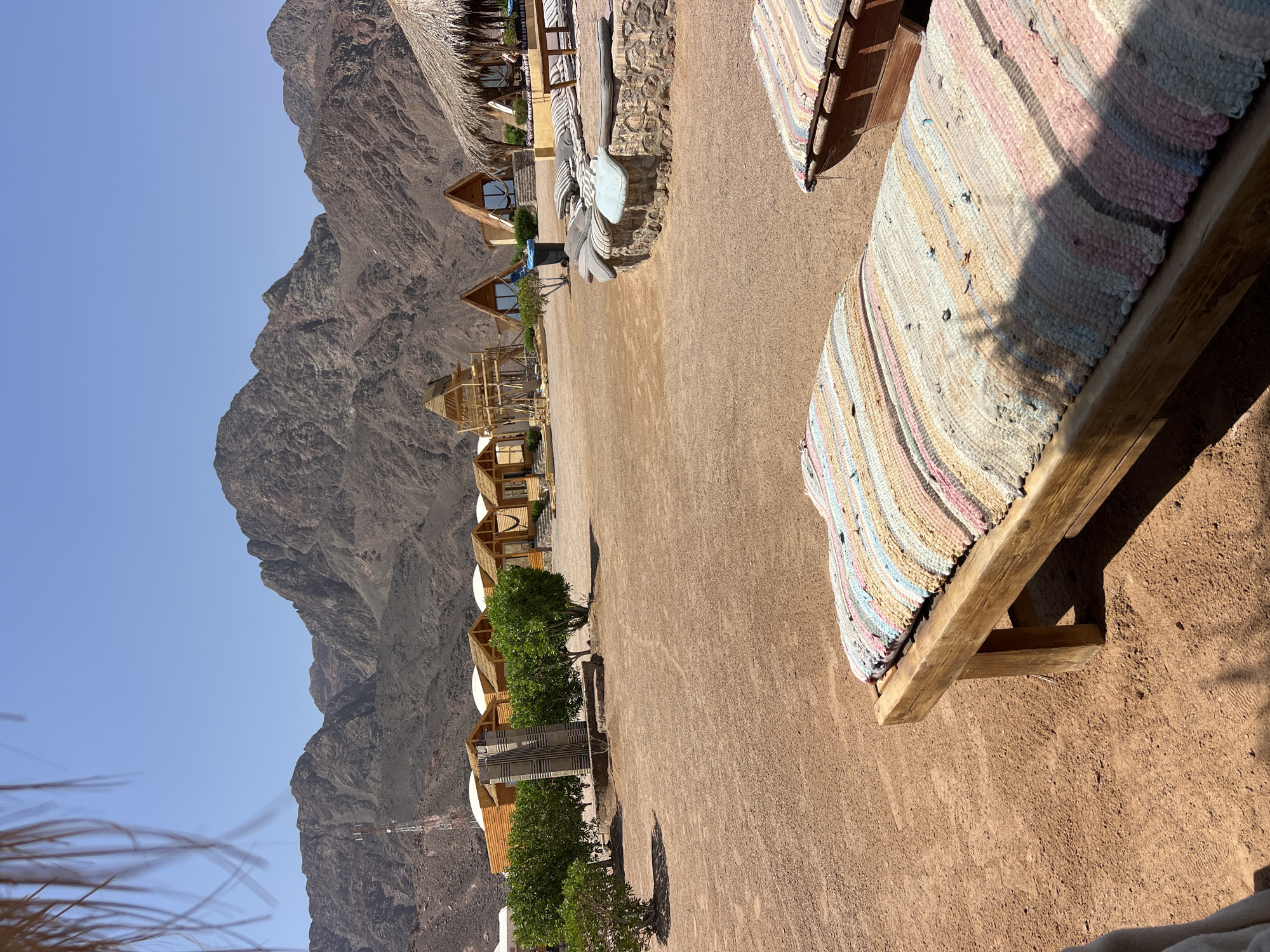 The image depicts an outdoor scene in a desert environment, likely a resort or campsite, in Nueiba, Egypt. In the foreground, two wooden benches with colorful striped cushions are positioned on reddish-brown dirt. Behind these benches, a low, curved stone wall is adorned with scattered cushions. Further back, a row of rustic wooden cabins with thatched roofs and triangular entrances are visible, nestled amongst sparse green trees. Some cabins have hammocks visible inside. To the left of the cabins, a tall, grey, monolithic structure stands against a backdrop of barren, rocky mountains under a clear blue sky. A telecommunications tower can be seen on the mountainside in the distance. The overall impression is one of a tranquil, minimalist desert retreat.