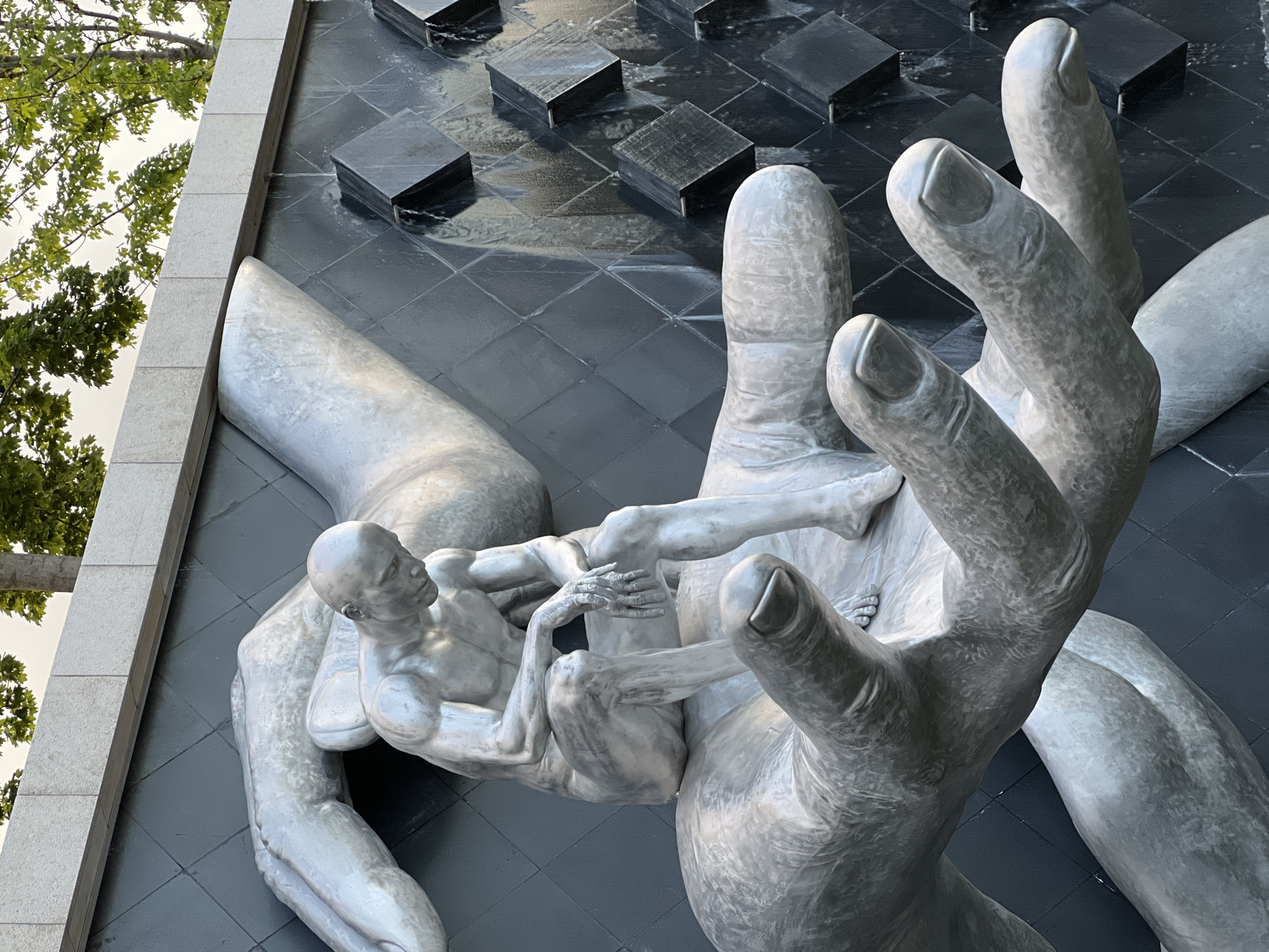 The image shows a metallic sculpture of a man in a pensive pose, resembling "The Thinker," seated on a giant hand. The sculpture is located in the city of Krasnodar, Russia, as part of a fountain or water feature. The man is depicted with visible musculature and is looking downwards. He is seated on a large, stylized hand which is itself supported by another, larger hand. Behind the sculpture, a dark, tiled wall serves as a backdrop, with water cascading down from cuboid structures embedded in the tiles. The scene is outdoors, with trees and their foliage visible at the top of the image, along with a section of a stone-like structure above the tiled wall, likely part of the building or architectural element supporting the fountain. The lighting suggests it is daytime, possibly overcast.