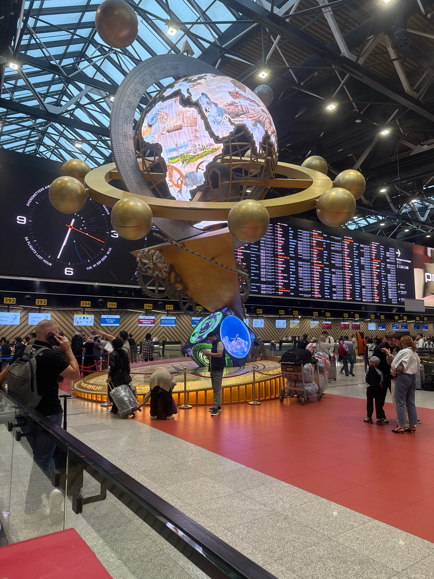 An indoor airport scene in Khimki, Russia, features a large, ornate clock with a globe in the center, surrounded by golden spheres. In the background, a digital departures board displays flight information. Several people are visible, including travelers with luggage, some queuing at check-in counters, and others conversing. The environment appears to be a busy terminal with polished floors and a modern ceiling structure.