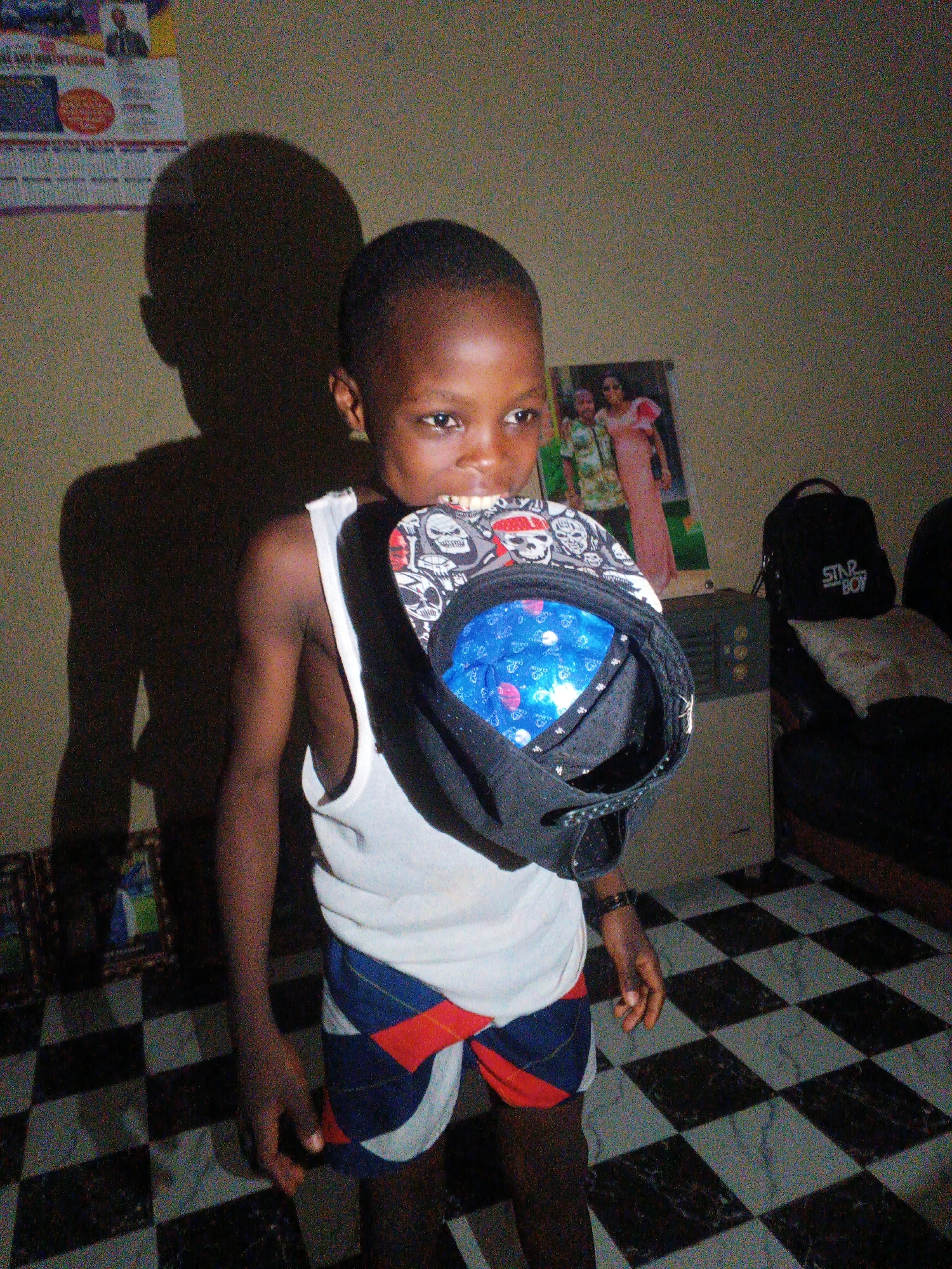 A young, dark-skinned boy stands in a room with a checkered black-and-white floor. He wears a white tank top and shorts with blue, red, and white diagonal patterns. He is holding a black baseball cap with skull designs on the brim in his mouth. The cap's interior has a blue pattern with white shapes.
Behind him is a beige wall with a shadow cast to the left. A poster with two women is visible, as well as a "Star Boy" branded black backpack on a dark couch. A small framed picture sits on the floor to the left, and a calendar hangs on the wall to the left of the shadow.
The location is likely indoors in Onitsha, Nigeria, given the subject's likely ethnicity and the overall style of the room.