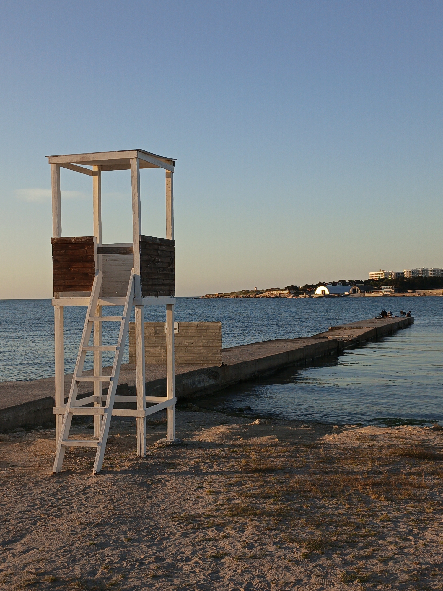 The image shows a lifeguard tower on a beach next to a pier in Sevastopol, Ukraine. The lifeguard tower is white with a wooden enclosure. The pier extends into the water, and people are sitting at the end. In the background, there is a building with a curved roof and other buildings along the coastline. The sky is a clear blue, and the sand on the beach appears coarse. The overall scene suggests a calm, late afternoon or early evening.