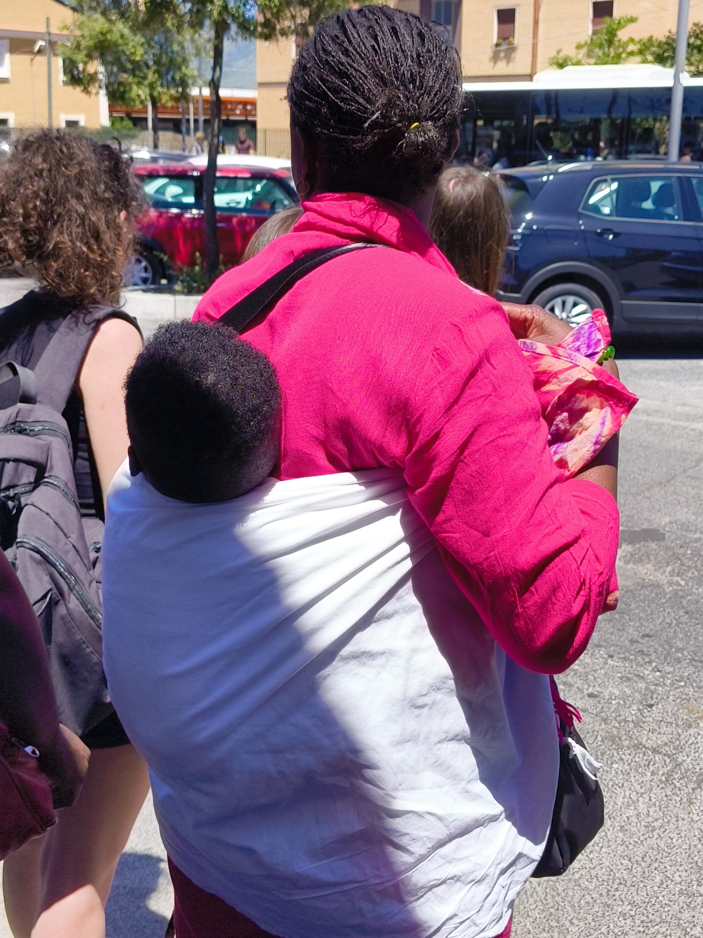 In the city of Fondi, Italy, on a sunny day, a woman with braided dark hair is seen from behind, wearing a vibrant pink top. She is carrying a baby wrapped in a white cloth against her chest. To the left, another person with curly dark hair is visible, wearing a grey backpack. In the background, a red car is parked, and a dark blue SUV is driving. Buildings with windows and trees are also present in the scene.