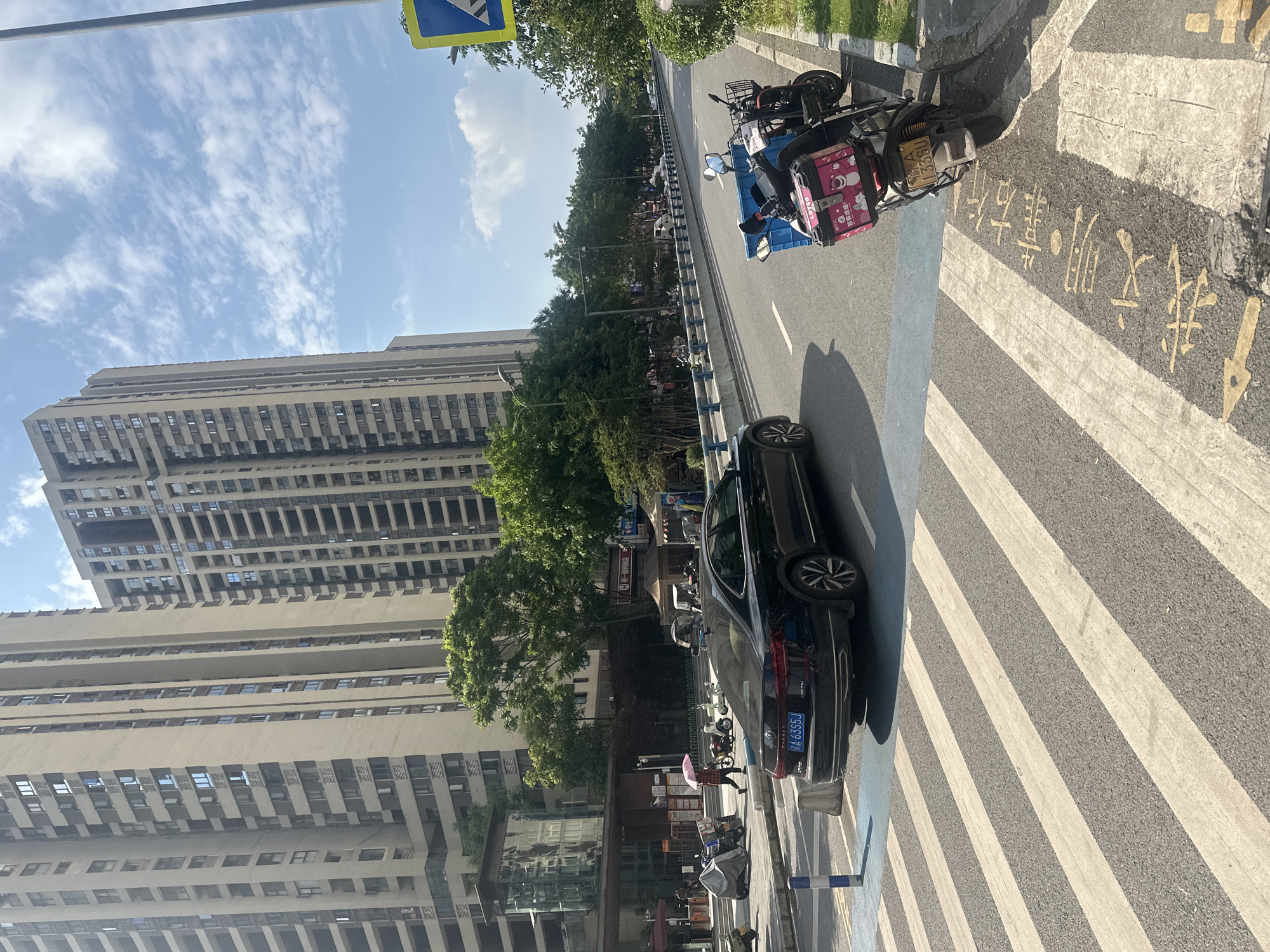 This image depicts a street scene in the Chongqing High-tech Zone, China. In the foreground, a pedestrian crossing with yellow and white stripes is visible. To the right of the crossing, a black sedan with a license plate reading "A 6355J" is parked. Further to the right, a scooter with a blue crate and a pink delivery box is parked on the sidewalk. The background features tall, modern apartment buildings and a row of green trees lining the street. A clear blue sky with scattered white clouds completes the scene. A pedestrian carrying a pink umbrella is walking on the sidewalk in the middle ground. The overall impression is of a bright, sunny day in an urban environment.