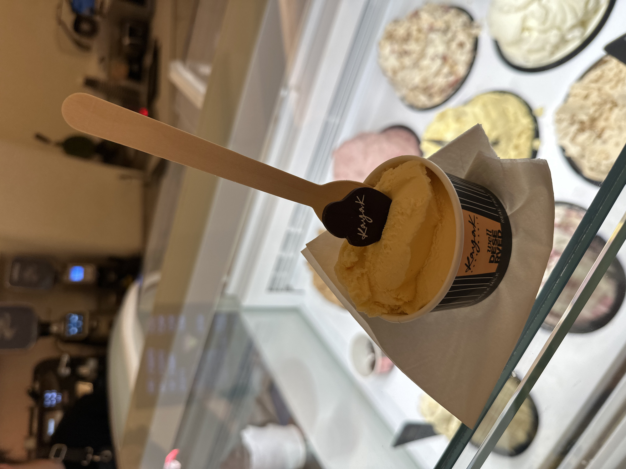 A close-up shot showcases a cup of yellow ice cream topped with a wooden spoon and a dark brown heart-shaped garnish with the word "Kayak" written in white cursive. The ice cream is in a black and orange patterned cup lined with a white paper napkin. The cup is placed on a glass display case, revealing various other flavors of ice cream in the background, including pink, white, and speckled varieties. The scene appears to be an ice cream shop in Mykonos, Greece, as suggested by the "Kayak" branding, a known ice cream parlor in the area. The lighting is ambient and the focus is on the ice cream, with the background blurred.