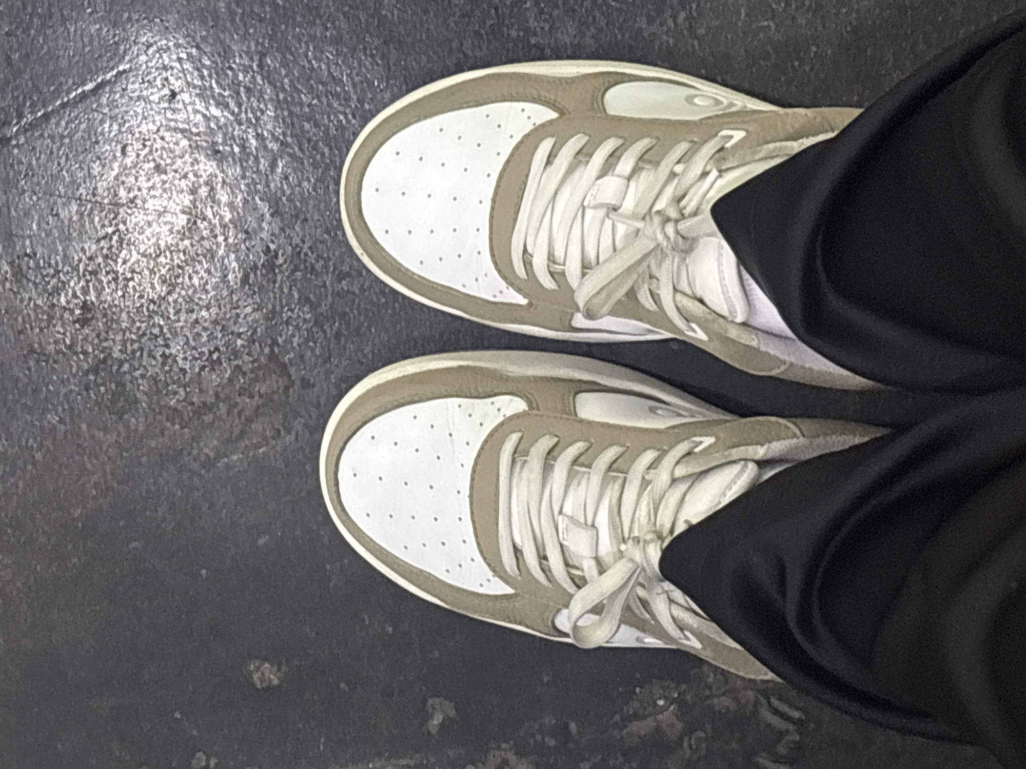 A top-down view of a pair of sneakers on a dark, possibly concrete floor. The sneakers are predominantly white with tan/beige accents around the edges and on the sides. The laces are white and tied. The right side of the frame is partially obscured by black fabric, likely pants, suggesting the perspective is from the person wearing the sneakers. The floor appears to be in an urban or indoor environment. No information indicates this image was taken in Buenavista, Mexico.