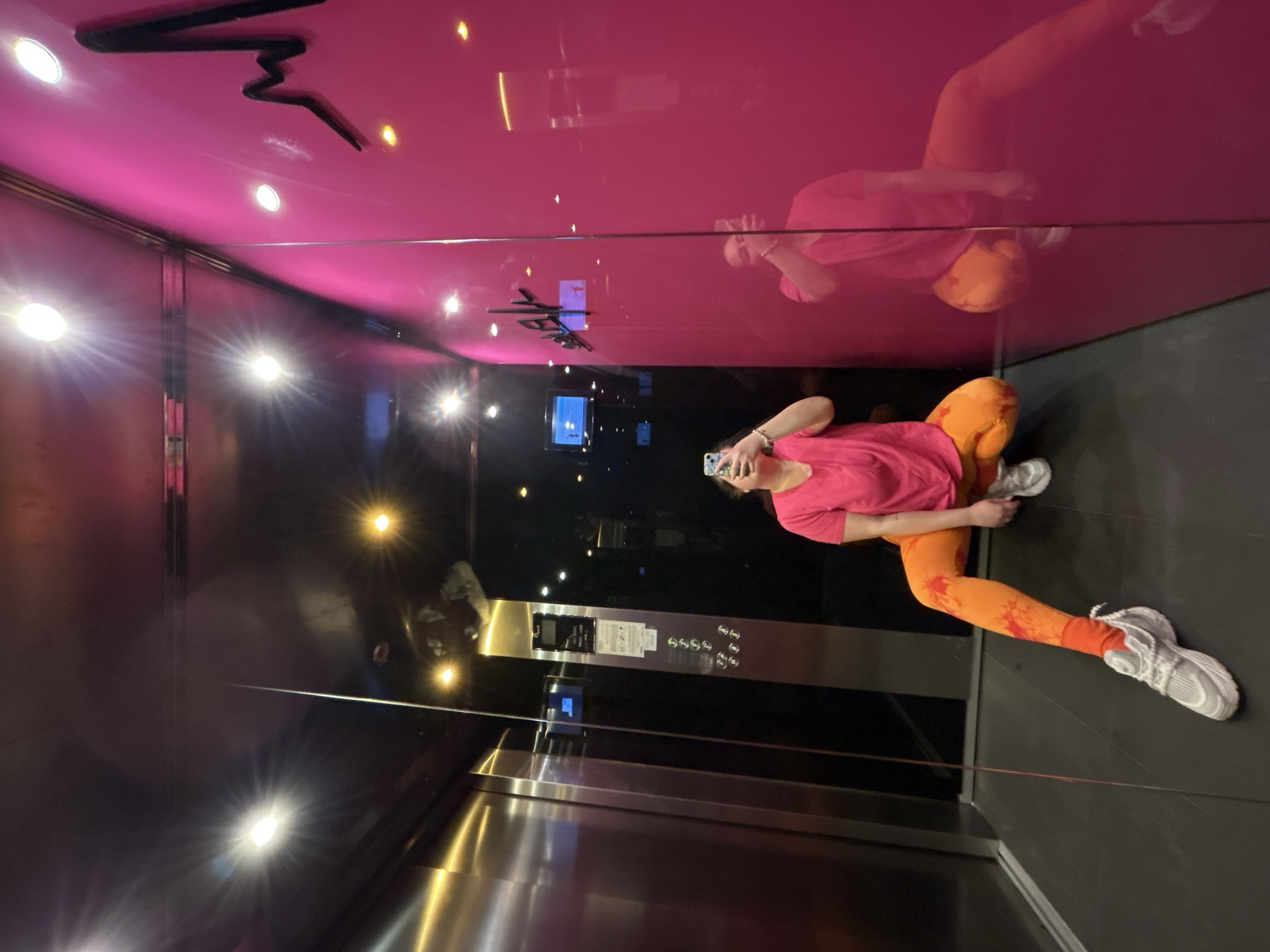 The photograph depicts an individual inside an elevator with reflective surfaces. The elevator's ceiling is bright pink with a black graphic design. The interior walls are a dark, reflective metal, reflecting the elevator lights and the person. The person is holding a phone, seemingly taking a picture of themselves. They are wearing a pink shirt and patterned orange leggings, paired with grey sneakers. The elevator's control panel is visible on the wall near the person. The location is identified as Plovdiv, Bulgaria, implied through additional information or context not visible in the image itself.
