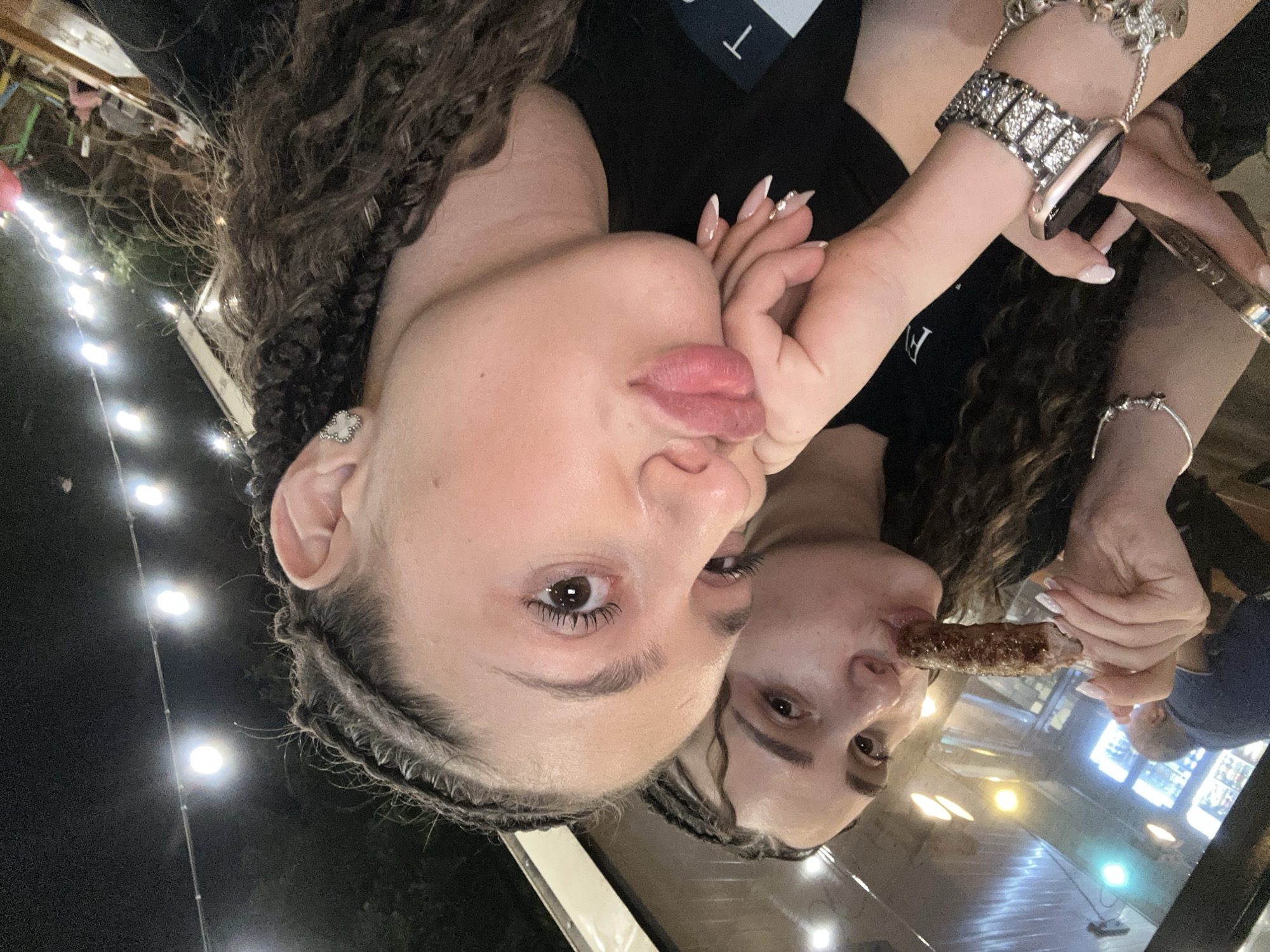 Two young women are taking a selfie outdoors at night. The woman in the foreground is leaning on her hand with her lips puckered. She is wearing a black t-shirt, a watch on her left wrist, and earrings shaped like clouds. The woman in the background is holding a piece of meat on a skewer to her mouth and is also wearing a black t-shirt. They are both in a dimly lit outdoor setting with string lights visible in the background. The location is identified as Gabrovo, Bulgaria.