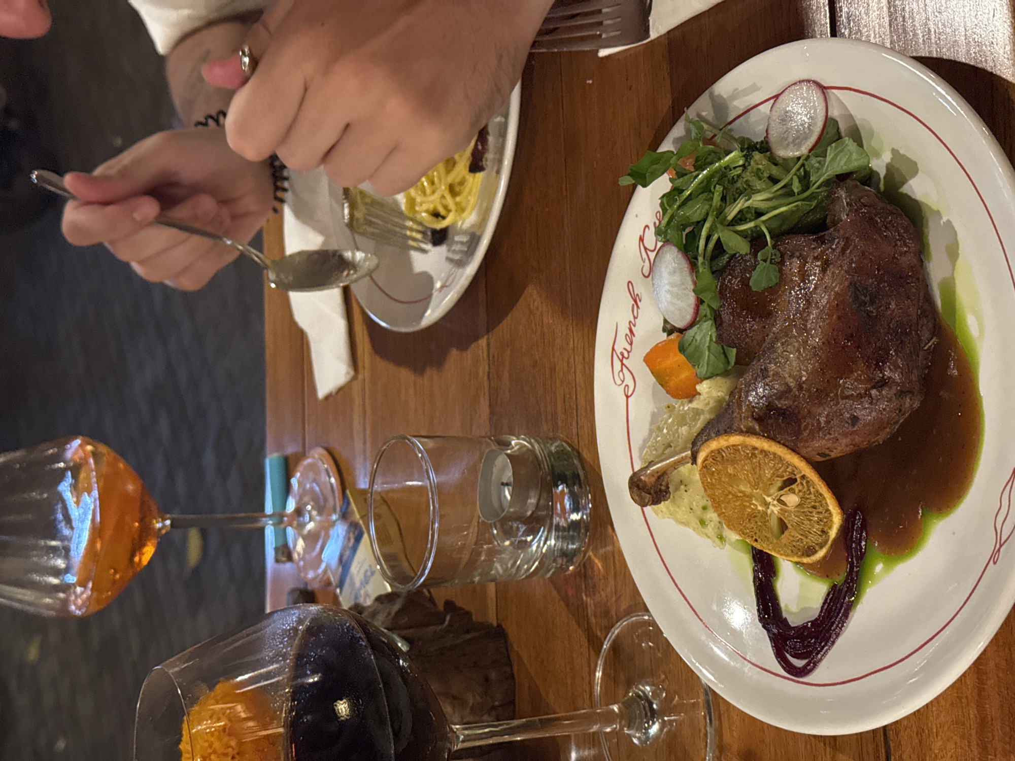 An overhead, medium shot captures a table at a restaurant, likely in Ubud, Indonesia, showcasing a meal and drinks. A white plate with a red rim is central, featuring a duck confit, mashed potatoes, a slice of dried orange, and a vibrant green sauce. To the left, a wine glass filled with red wine and another with an orange aperitif stand prominently. A small glass votive with a lit tea light sits nearby. In the background, a person's hands are visible, one holding a fork over a plate of pasta, and the other gesturing with a ringed finger. The table's wooden surface is visible throughout the scene.