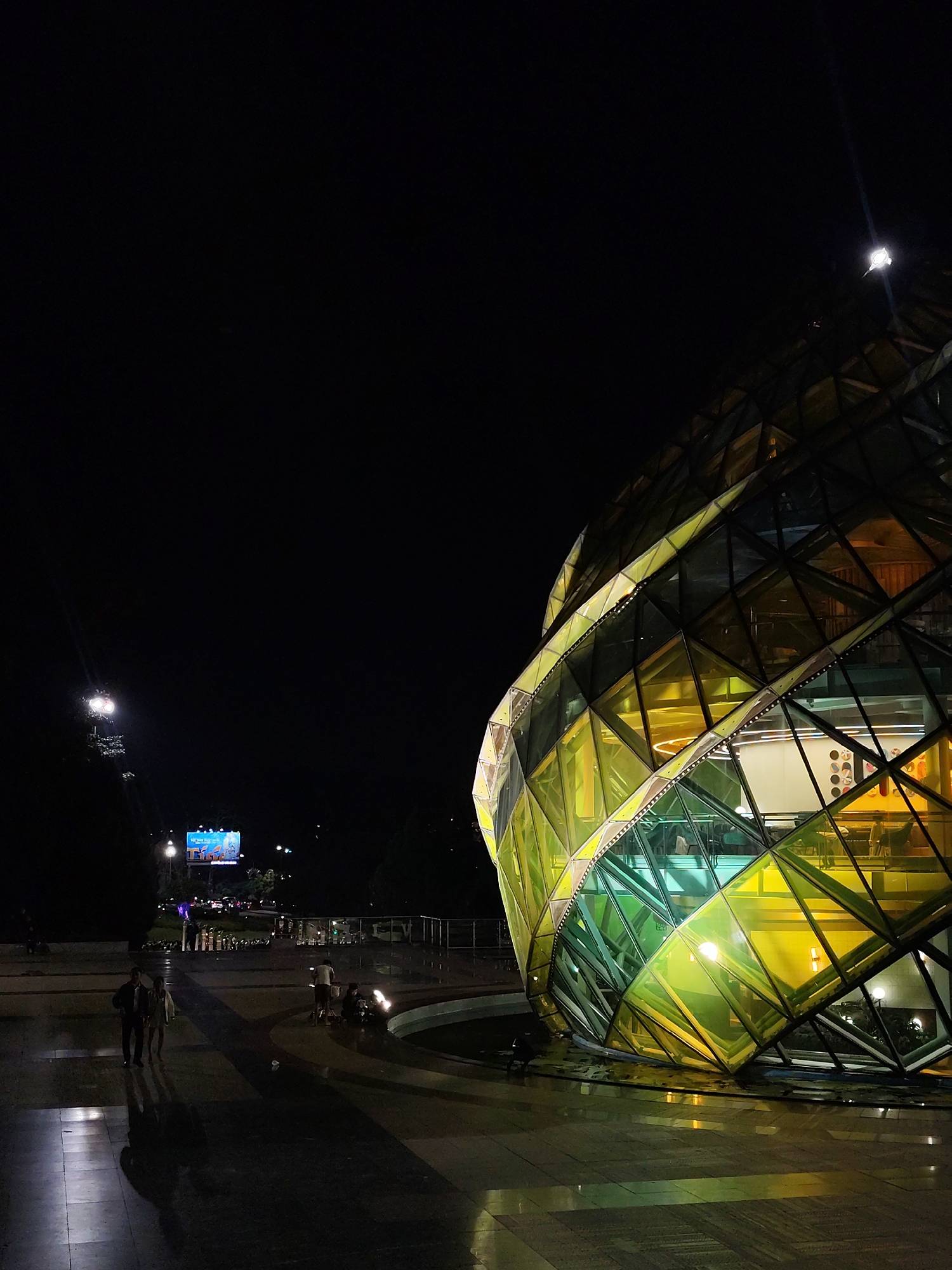The image depicts a night scene in Da Lat, Vietnam, featuring a distinctive, multifaceted glass building with a geometric, almost crystalline design, illuminated with yellow and green lights. The building appears to be part of a public space or entertainment venue, as suggested by the presence of people walking on the paved ground and what seems to be a fountain or water feature in the foreground. The building's reflective glass panels create a vibrant, almost otherworldly appearance against the dark night sky. In the background, a large billboard displaying advertisements is visible, along with streetlights and distant lights indicating an urban environment. The overall atmosphere is one of modern architecture and nighttime activity.