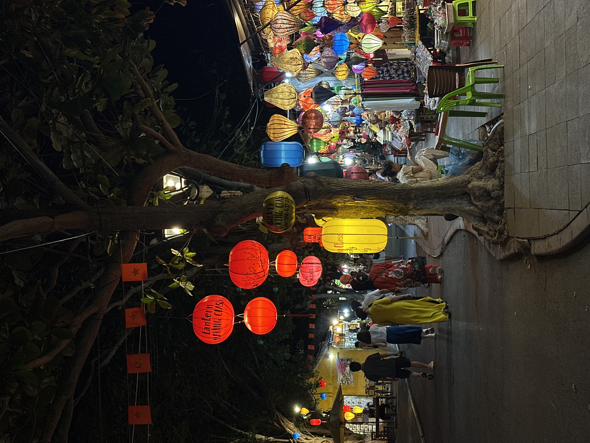 This is a photograph taken at night on a street in Hoi An, Vietnam, likely near the Ancient Town, renowned for its vibrant lantern displays. The scene is dominated by numerous colorful lanterns of various shapes and sizes hanging from trees and stalls. Red, yellow, blue, and green lanterns are visible, illuminating the street and creating a festive atmosphere.
In the foreground, a paved walkway curves to the right. Several people are walking away from the camera on the left side of the street, including a man in a dark shirt and shorts, a woman in a white top and blue skirt, a woman in a long yellow dress, and a woman in a red garment with a floral pattern.
Further down the street, stalls are set up selling a variety of goods, predominantly more lanterns and textiles. A woman is seated on a green plastic chair to the right of a large tree, appearing to be engaged in an activity. Trees with dark bark and green leaves are prominent, with branches extending over the street. Small red flags with yellow stars are strung across some of the trees in the background. The lighting is dim, with the lanterns providing the primary sources of illumination, casting a warm glow on the surroundings. The overall impression is one of a lively night market or festival.
