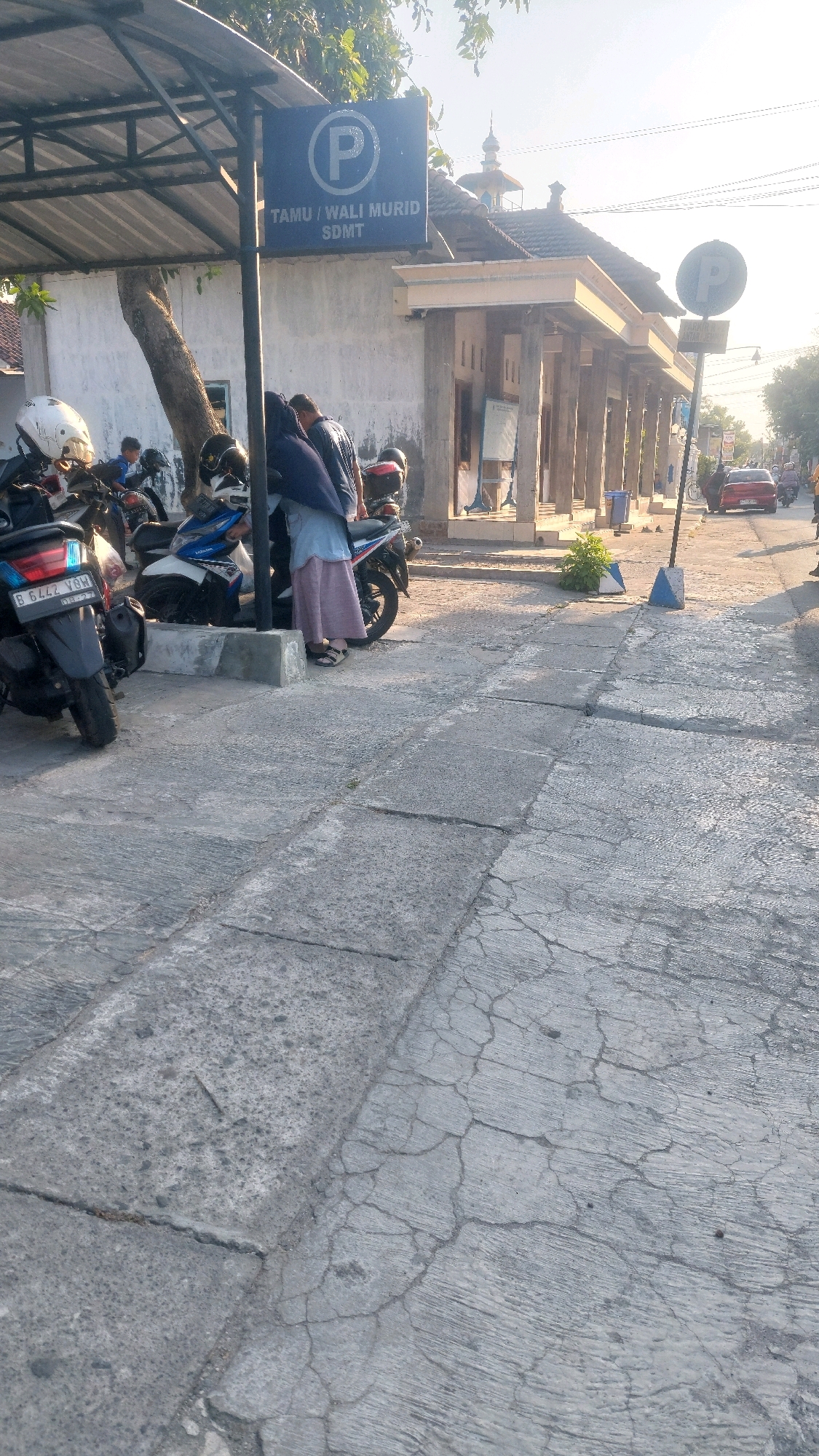 A parking sign for guests and parents of SDMT (Sekolah Dasar Muhammadiyah Timbul) is visible in the city of Ronowijayan, Indonesia. The sign is blue with a white circle and a white "P" inside, with text below reading "TAMU / WALI MURID SDMT". Several motorcycles are parked in the area, suggesting it is a school parking lot. A few people are standing near the motorcycles, and some appear to be interacting. The environment is an outdoor paved area, possibly a sidewalk or parking lot, with some vegetation and a building in the background. The lighting suggests it is daytime, with sunlight casting shadows. A red car is visible on a road in the distance. The pavement is cracked and worn in some areas.
