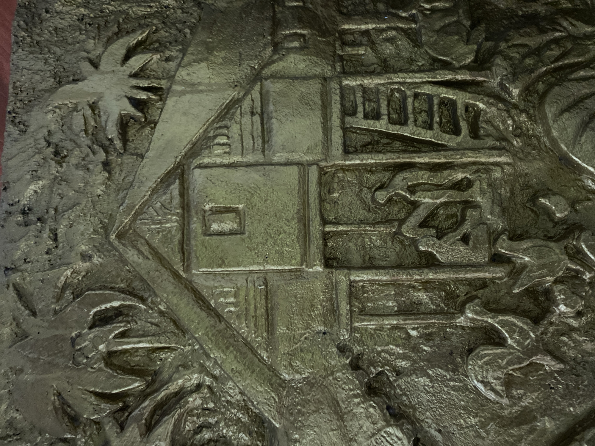 This is a close-up, overhead shot of a relief sculpture, likely made of metal and painted a dull, yellowish-green. The sculpture depicts a rural scene. In the center is a stylized, stilted house with a peaked roof. There is a small, square window on the upper part of the house. In front of the house, beneath the overhang, three figures are depicted in low relief, appearing to be engaged in some activity. One figure is seated, while the other two are standing behind them. To the right of the house is a ladder. The foreground of the sculpture features several animal figures, which appear to be chickens or similar fowl. Palm fronds are visible on the left and right sides, suggesting a tropical or subtropical setting. The overall texture of the sculpture is rough and uneven, with deep grooves and raised areas contributing to the three-dimensional effect. The lighting is dim and appears to be coming from above and slightly to the right, casting shadows that enhance the relief.