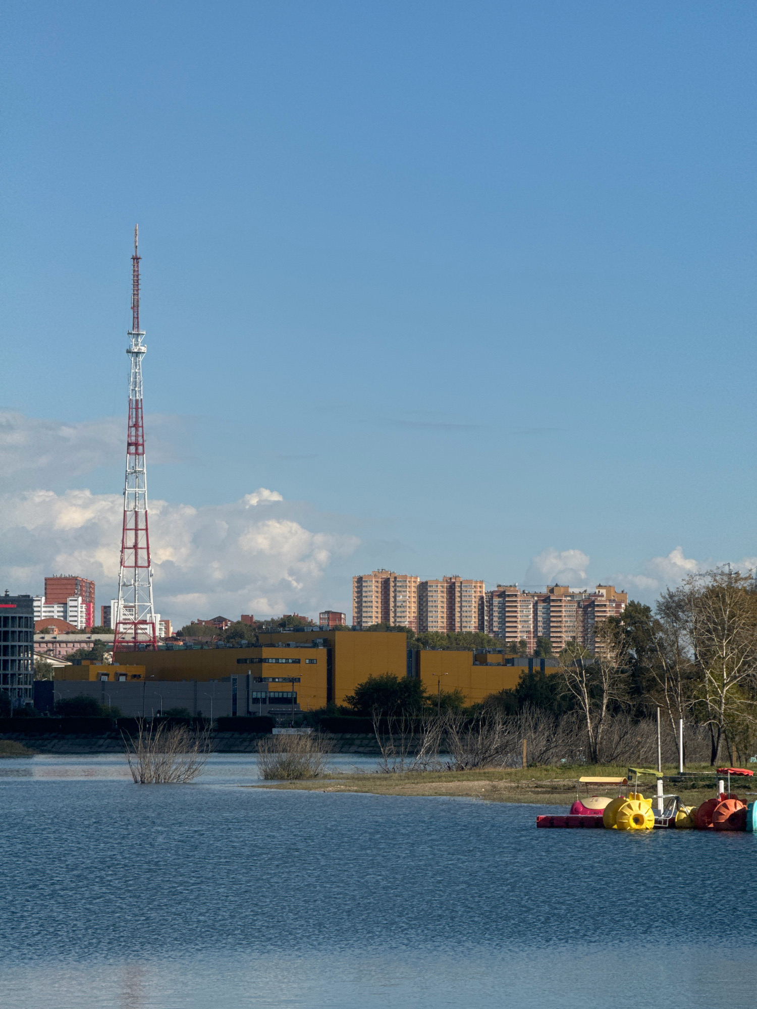The image depicts a waterfront scene in Irkutsk, Russia, featuring a television tower and apartment buildings. In the foreground, a tranquil blue body of water stretches across the frame, with several colorful paddle boats docked along the right edge. The water exhibits gentle ripples, indicating a calm day.
The middle ground reveals a sandy embankment with sparse vegetation and bare trees, suggesting an autumn or early spring setting. Beyond the embankment, a low-lying yellow building with dark windows is visible, likely a commercial or industrial structure. Further in the background, a towering television tower with red and white crossbeams dominates the skyline on the left side. Behind the yellow building and the tower, a cluster of multi-story apartment buildings, mostly in shades of red and orange, are situated. The sky is a clear, bright blue with scattered white clouds. The scene is bathed in bright sunlight, casting subtle shadows and highlighting the textures of the water and the buildings. No people are visible in the frame.