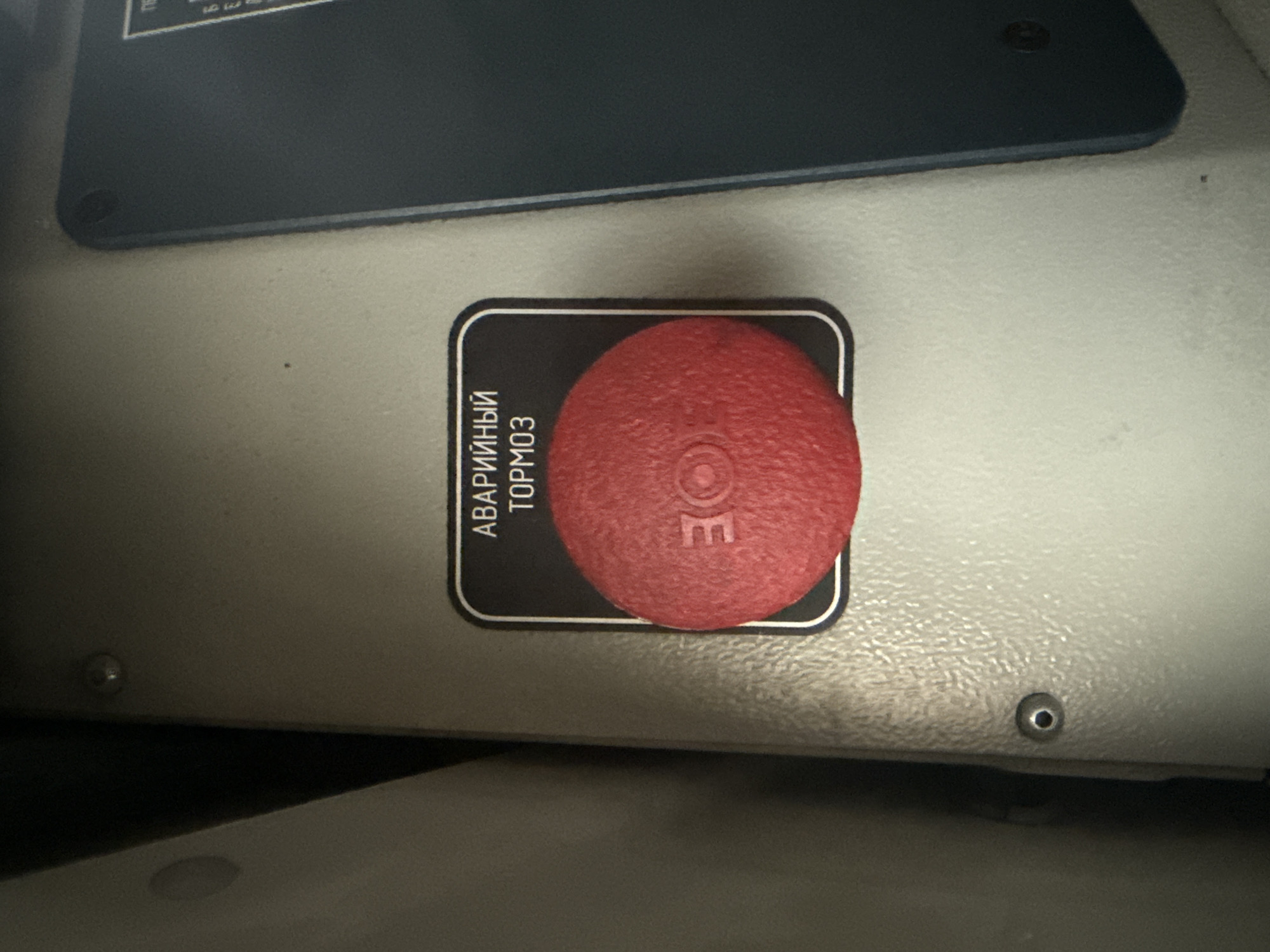 The image shows a close-up of a red emergency stop button mounted on a gray panel. The button is round and has the Cyrillic text "АВАРИЙНЫЙ ТОРМОЗ" (Emergency Brake) above it. There is a circular indentation on the button itself. The panel appears to be part of some industrial or transportation equipment. The background is dark and out of focus. There are no people or signs of activity in the image. The provided image does not contain any information or context related to the city of Verkhnezeysk, Russia.
