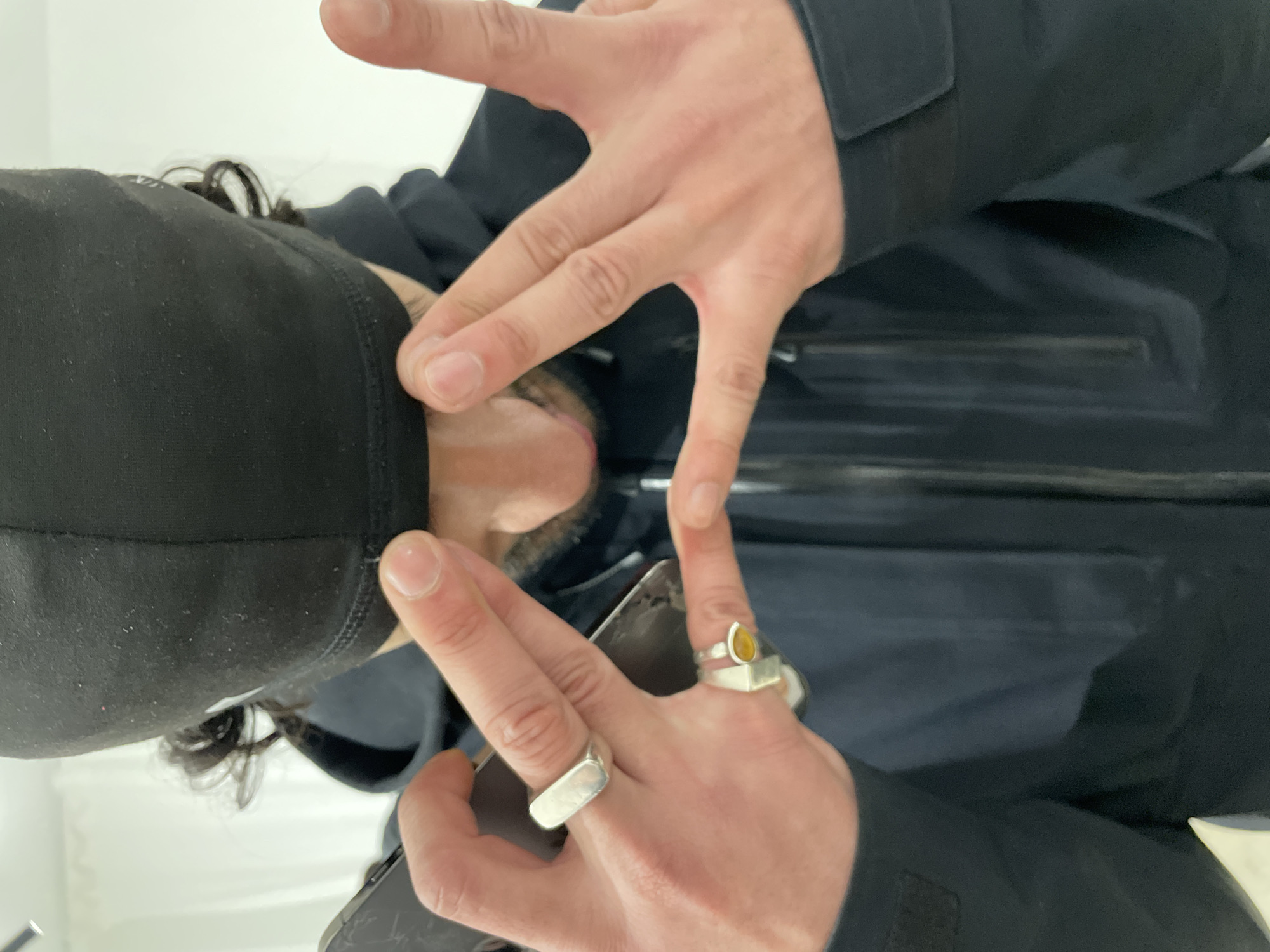 A person, gender indeterminate but appearing masculine, is depicted in a close-up, high-angle view. Their face is largely obscured by a black balaclava or beanie pulled down to the upper lip, with dark, curly hair visible emerging from beneath. A beard or stubble covers the chin and jawline, and reddish discoloration is apparent around the nostrils. The subject wears a dark hooded jacket. The left hand holds a dark smartphone with a cracked screen; a rectangular silver ring is on the ring finger and a silver ring with an amber-colored stone is on the middle finger. Both hands are raised in front of the lower face, with fingers spread to form a 'W' or 'V' gesture. The background consists of a plain white wall, suggesting an indoor setting. The specific location within Paris, France, cannot be determined from the provided visual information.