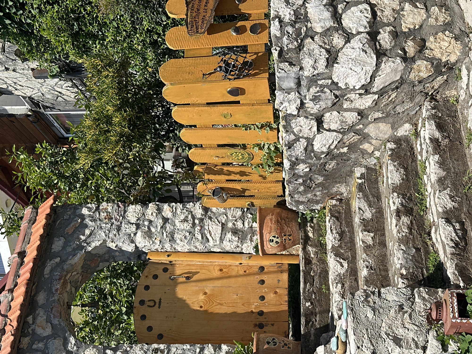This is an exterior shot of a stone stairway leading upwards, possibly in Budva, Montenegro. The stairway is constructed of gray stone with visible mortar joints. Along the right side of the steps is a wall made of similar stone construction. A wooden gate is visible at the bottom of the stairs, constructed of vertically arranged planks. The gate appears to be a light brown color. Next to the gate, there is a stone archway with a red tiled roof. The archway is surrounded by green foliage. A wooden fence is placed above the stone wall, constructed of rounded planks painted a golden yellow. The fence features decorative objects, including a metal insect sculpture. The environment is overgrown with greenery. A building can be seen in the background behind the foliage.