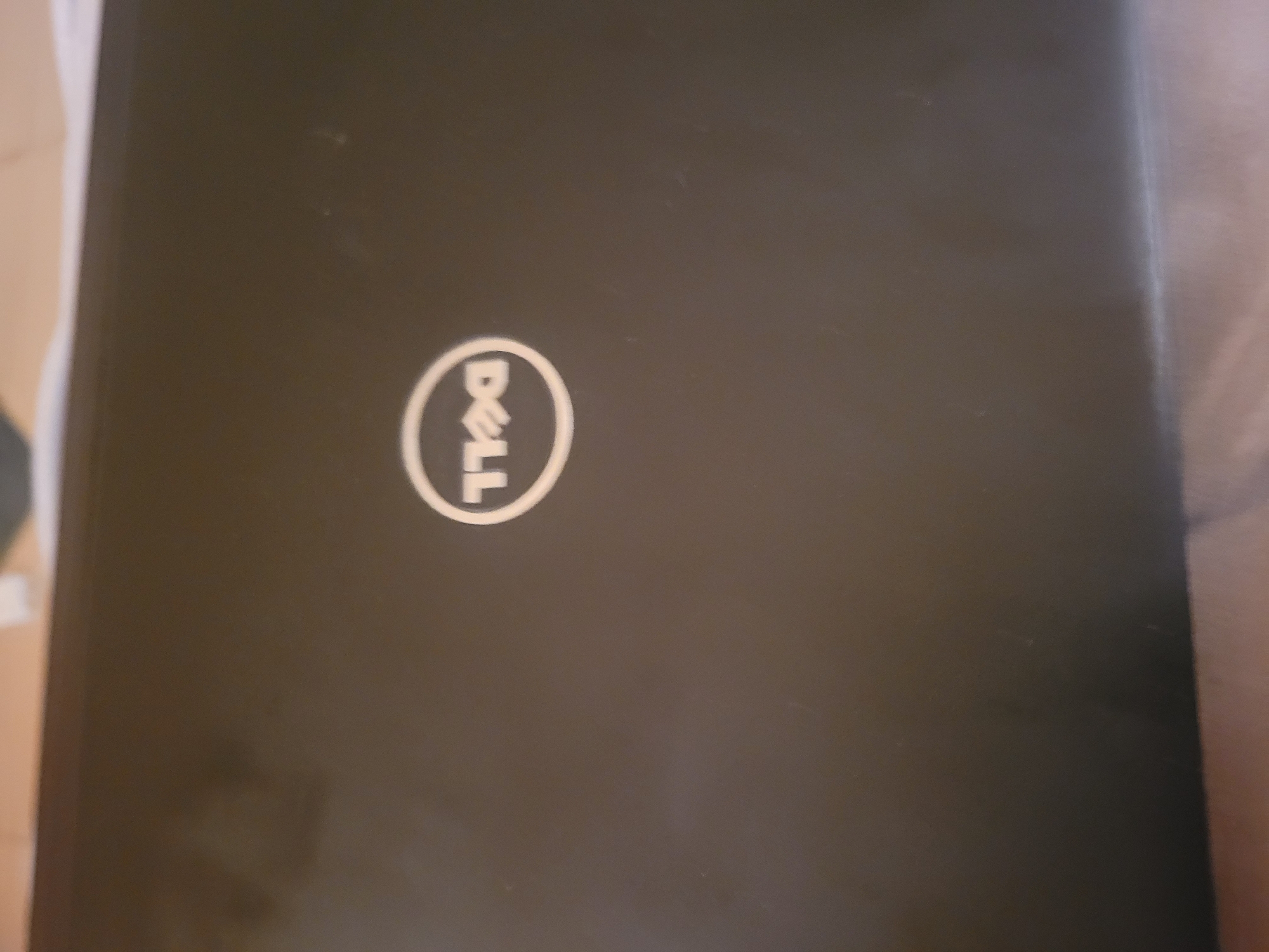 A closed black Dell laptop is shown from a top-down perspective. The Dell logo, a white circle with the company's name inside, is centered on the lid. The background consists of a blurred surface, possibly a table or desk, with a soft, warm tone. The lighting is diffuse, creating a slightly soft focus on the laptop's surface, which appears to be matte. There are no people or other objects visible in the image. The scene is static and indoor.