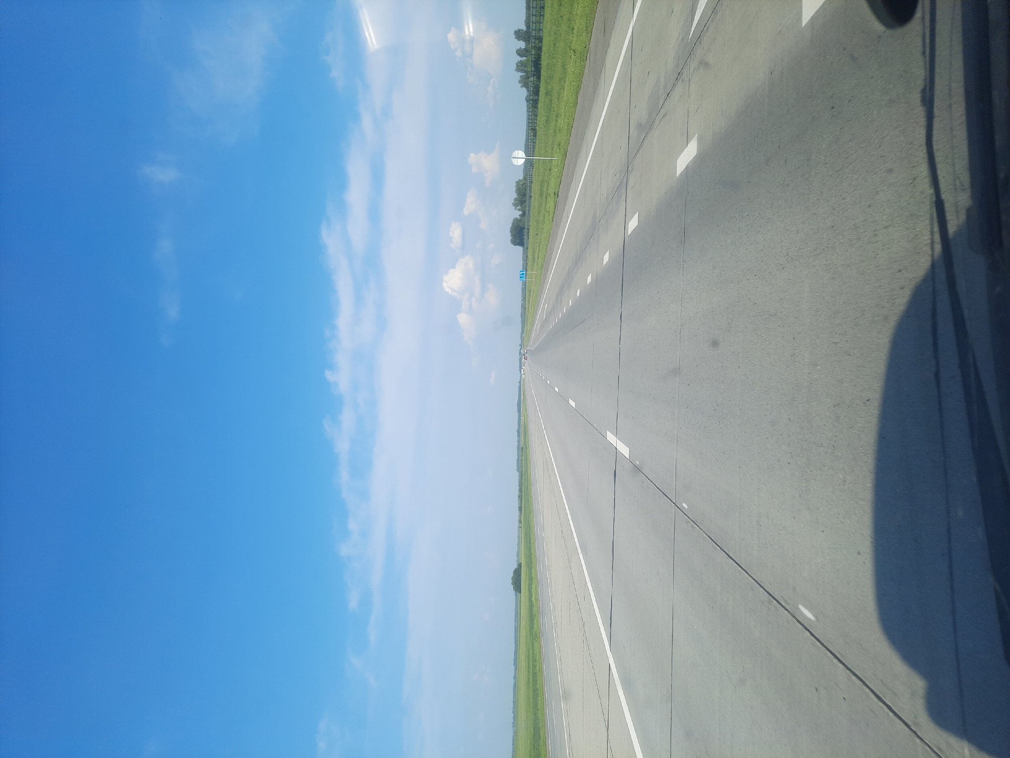 The image depicts a wide, straight asphalt road stretching into the distance under a bright blue sky with scattered white clouds. The road is divided by white lane markings. To the left and right of the road are flat, green fields, with a fence line visible in the distance on the right. There are a few scattered trees and some distant buildings or structures. On the right side of the road, a round white sign with a blue symbol and a smaller white sign with a blue symbol are visible. A dark shadow, likely from a vehicle, is cast across the bottom foreground of the image. The scene appears to be located on the outskirts of Pokrovka, Russia, given the flat terrain, open fields, and the style of road markings and signage.