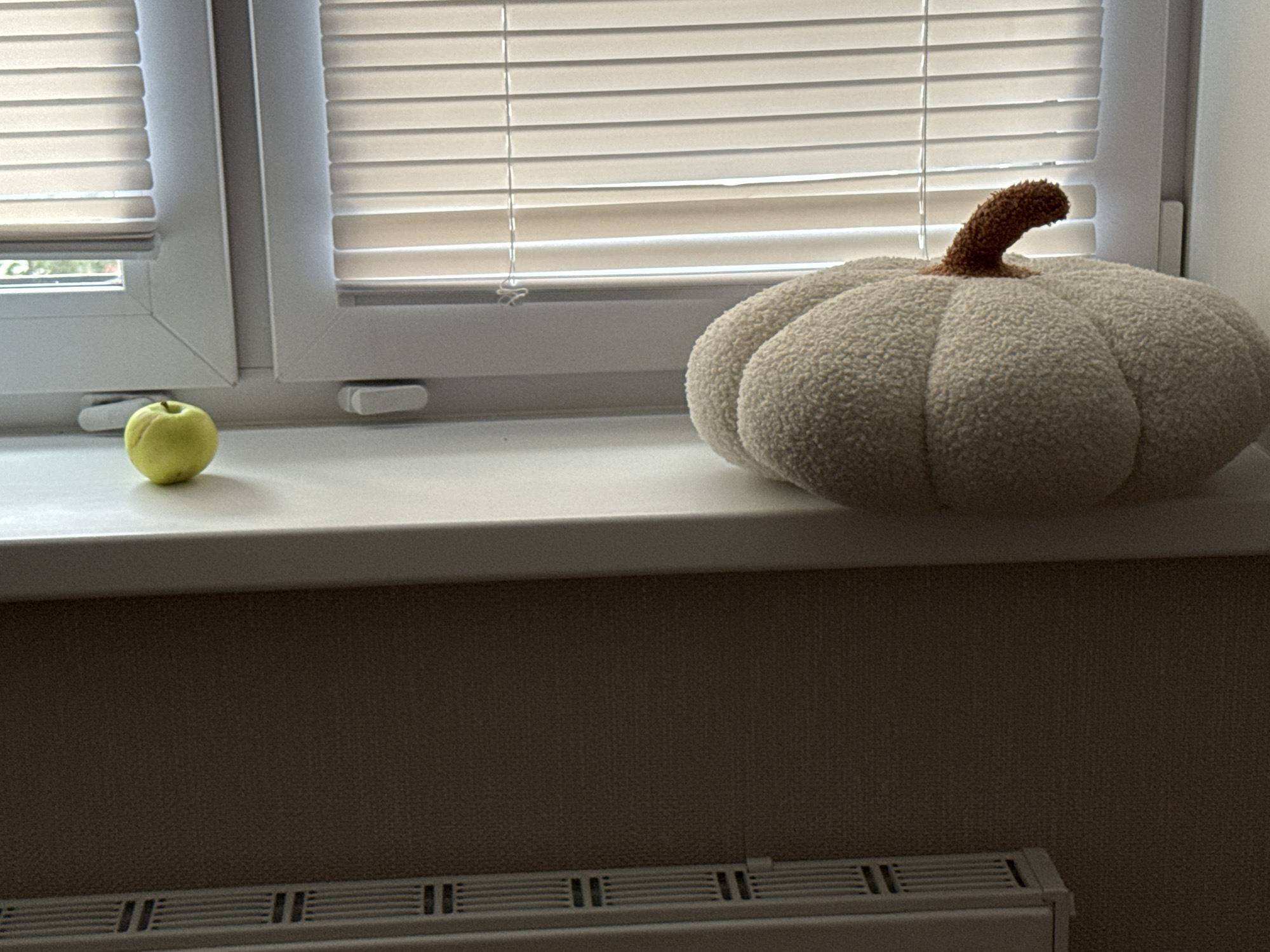 A light green apple sits on a white windowsill next to a plush, cream-colored pumpkin-shaped pillow. The windowsill is in front of horizontal blinds that cover a window, allowing diffused light to enter the room. Below the windowsill, a brown textured wall is visible, and at the bottom of the image, a white radiator with horizontal vents can be seen. The scene is indoors, likely in a residential setting, with no people present. The overall lighting suggests it is daytime.