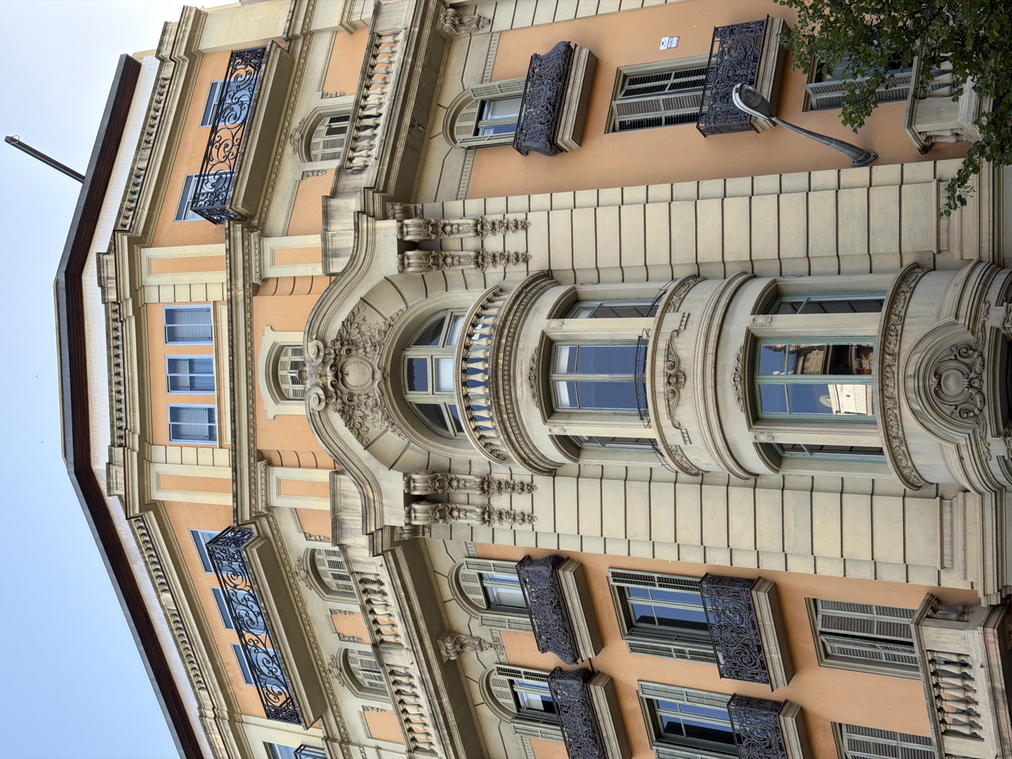 The image shows the ornate facade of a building in Barcelona, Spain, likely located in the Eixample district, known for its modernist architecture. The building features a salmon-pink exterior with cream-colored decorative moldings and balconies. Several windows with wooden shutters are visible, some open and some closed. Ornate wrought-iron balconies, characteristic of the era, adorn various levels of the building. A prominent curved bay window with decorative stonework and a wrought-iron balustrade stands out in the center of the facade. A street lamp is affixed to the exterior wall on the right side of the building, extending outwards. Lush green foliage from a tree partially obscures the lower right portion of the building. The sky above is a clear, pale blue. No people are visible in the image.