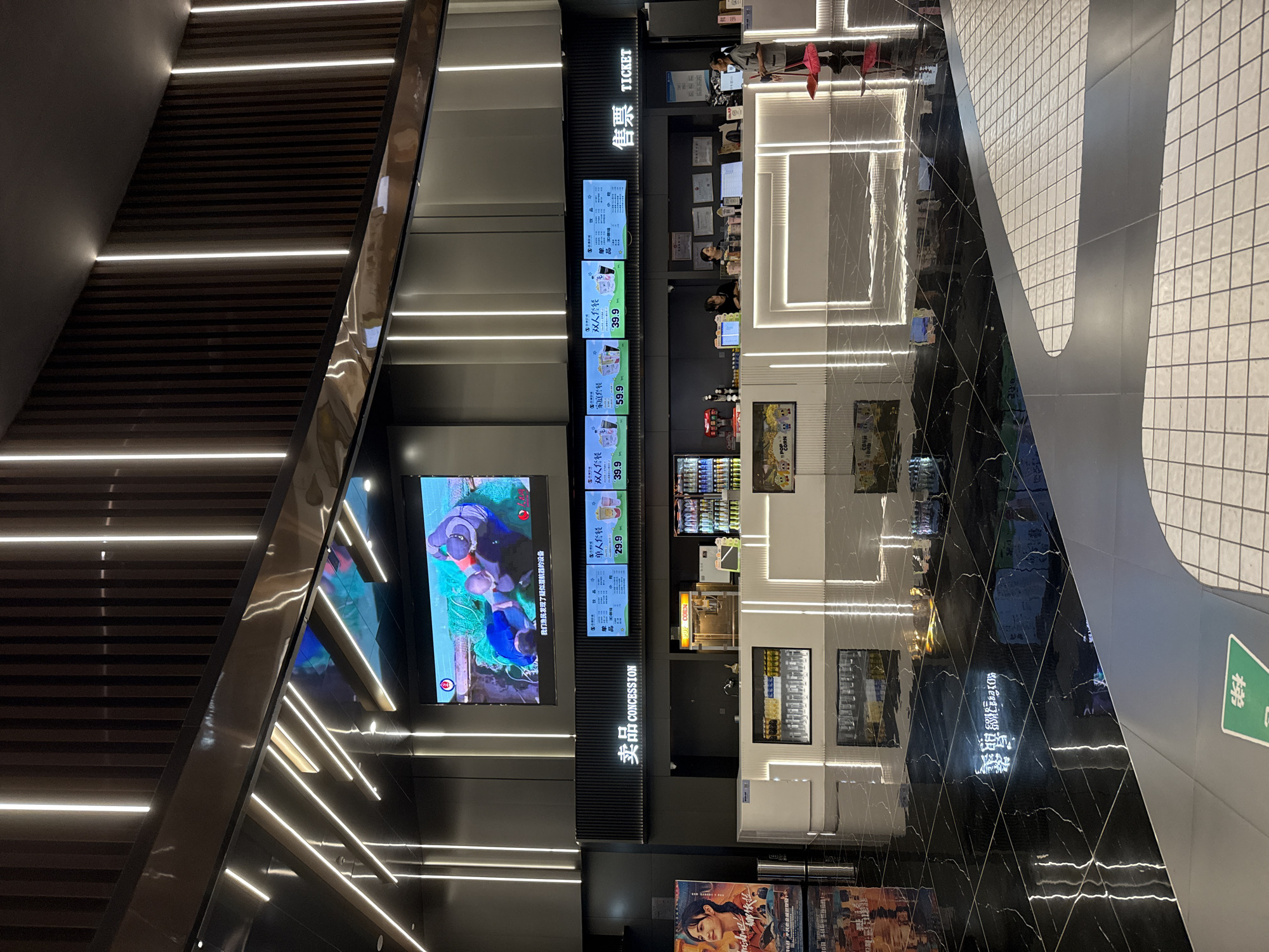 The image depicts the interior of a modern cinema lobby in Huizhou, China. In the center, a concession stand with a bright, illuminated counter is visible, offering various beverages and snacks. Above the counter, digital menu boards display prices and item names in Chinese characters. A large screen displaying movie trailers or promotions hangs prominently at the top of the scene. The floor is polished black marble, reflecting the ambient lighting from the numerous vertical LED strips that adorn the walls and ceiling, creating a sleek and contemporary atmosphere. Movie posters are displayed on the left side of the frame. To the right, a cleaner is seen sweeping the floor with a red broom and dustpan. Several people are interacting at the concession stand, suggesting a normal operational day at the cinema.