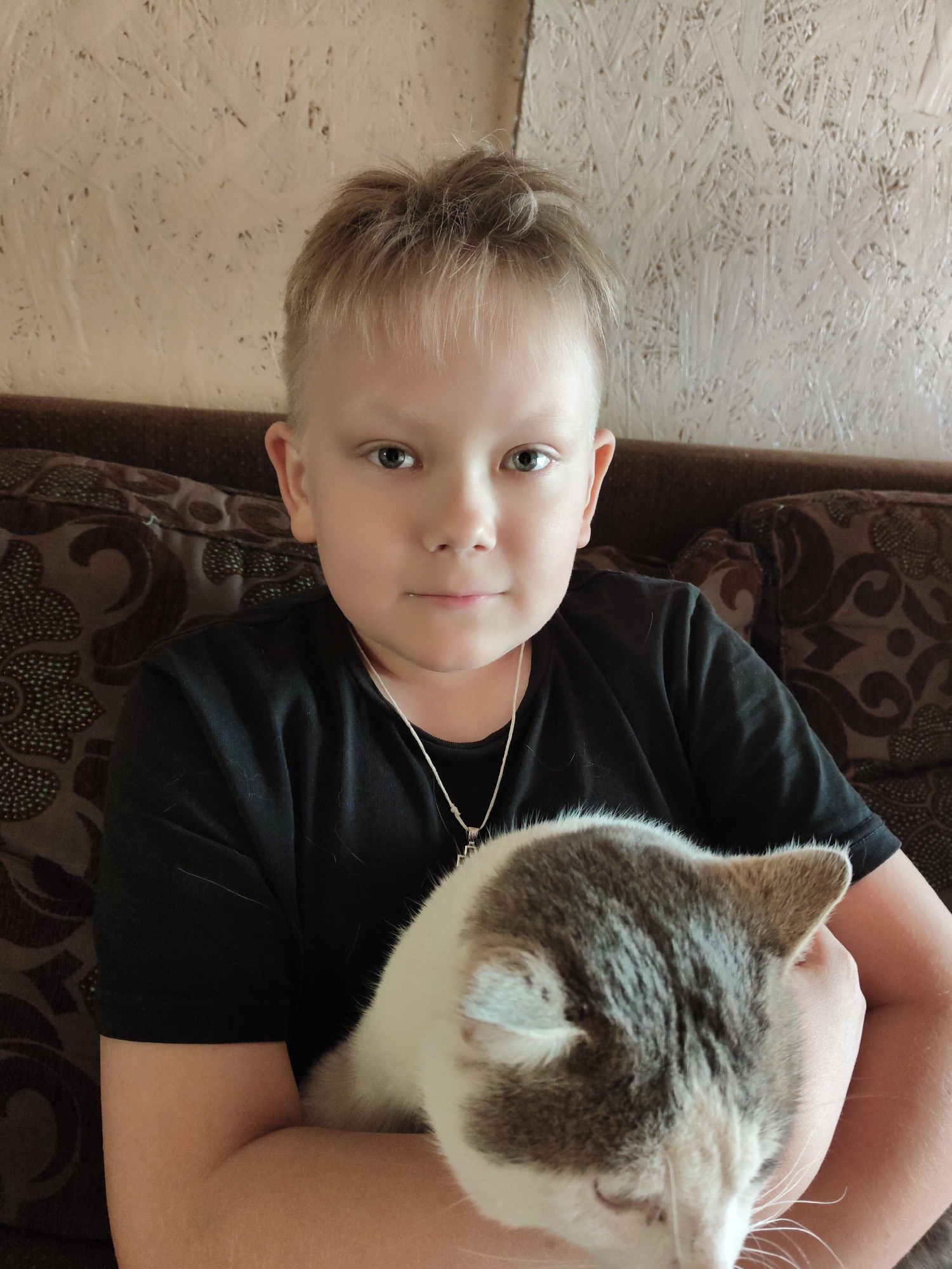 A medium shot depicts a fair-skinned child with light blonde hair, wearing a black t-shirt and a silver necklace with a cross pendant. The child is holding a gray and white cat in their arms. They are sitting on a brown patterned couch, in front of a textured beige wall. The location appears to be indoors. The location is in the city of Unknown, Unknown.