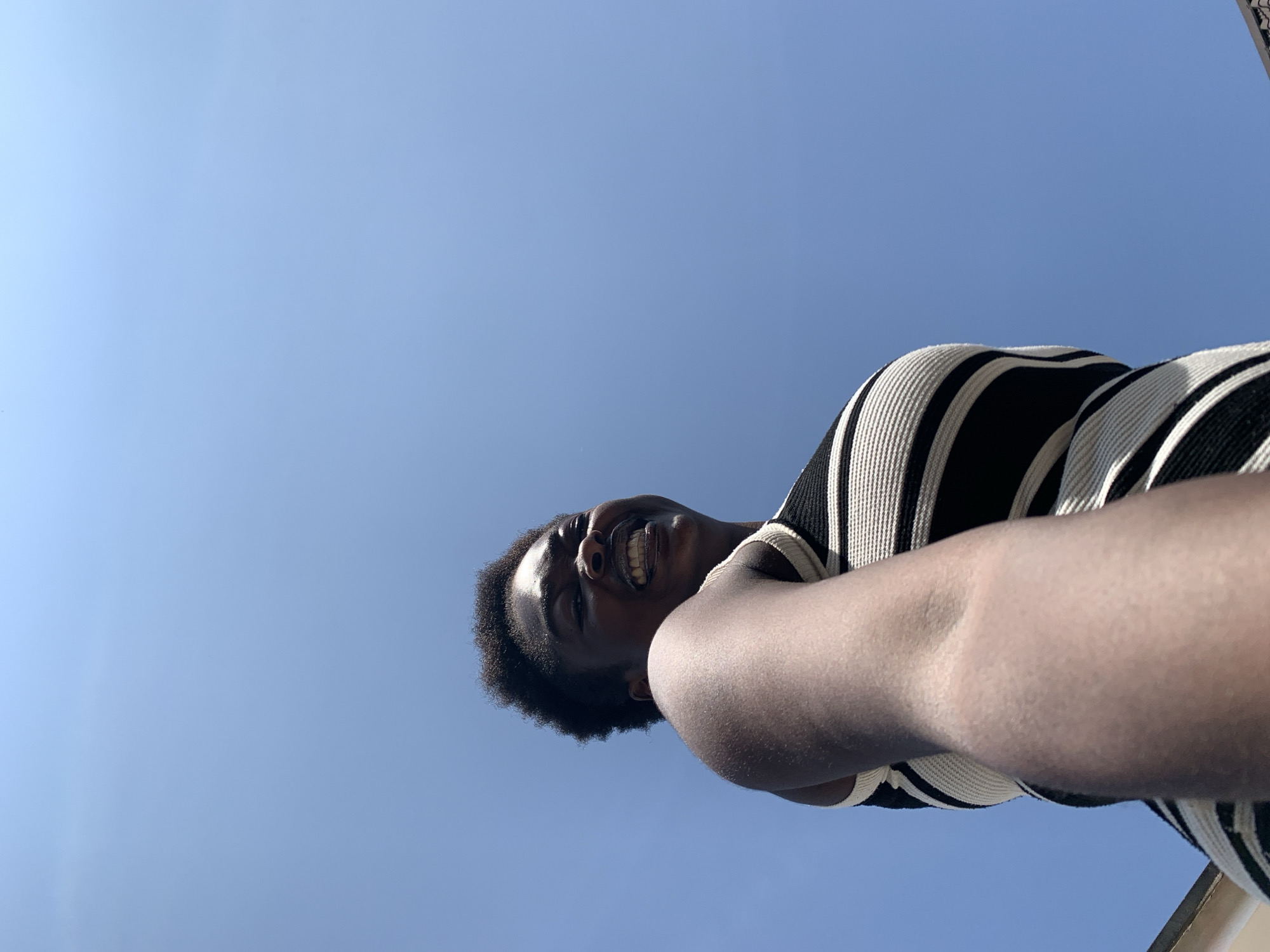 A low-angle, close-up, outdoor, daytime shot captures a Black woman with short, dark, curly hair and a bright smile against a clear, bright blue sky. She is wearing a black and white striped, sleeveless top. Her left arm extends into the foreground, and she is looking back over her left shoulder towards the camera with a joyful expression. The image appears to be a selfie. There are no visible buildings or other contextual environmental details that would indicate a specific location within Gura Top, Nigeria.