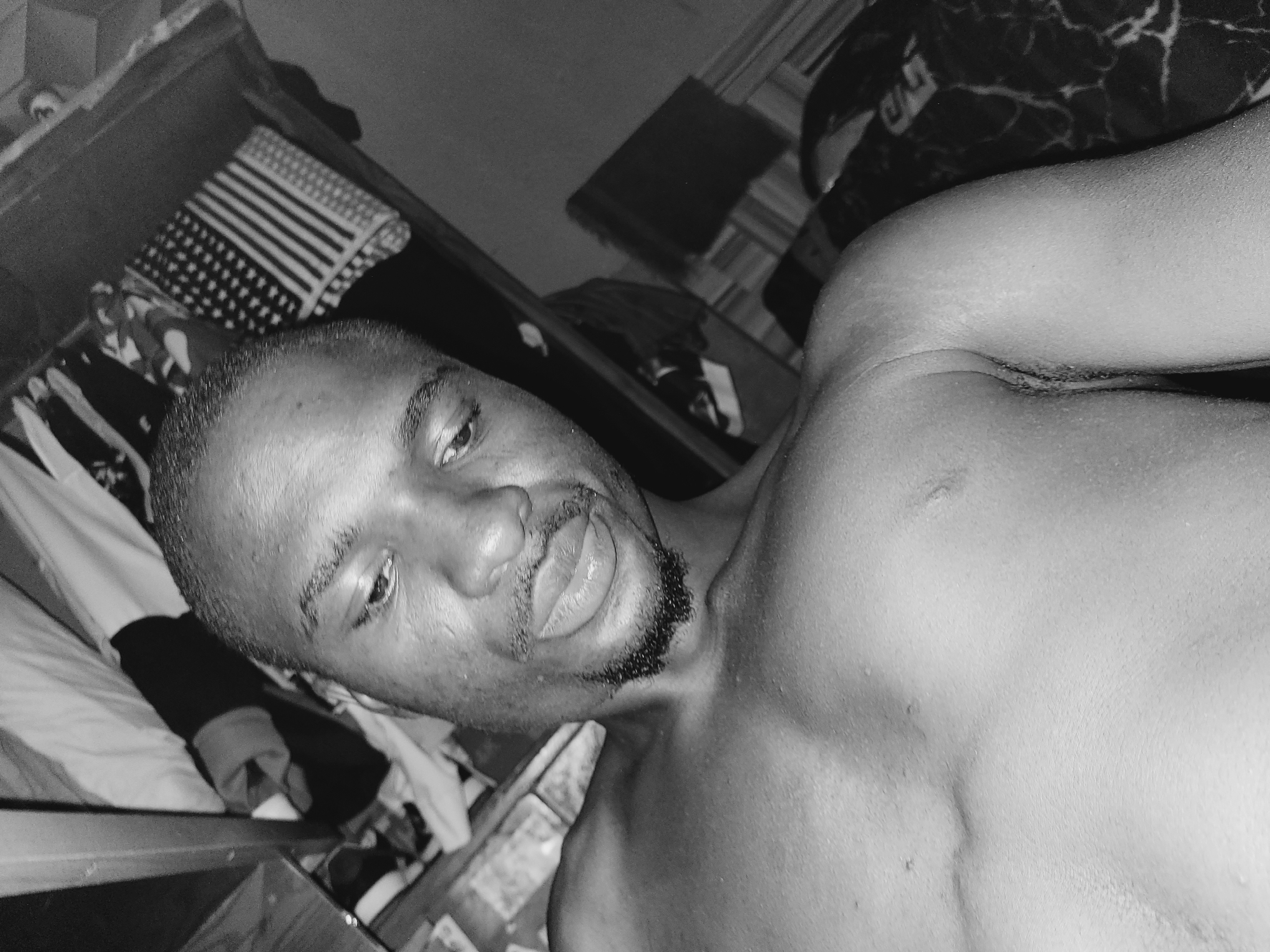 The image is a black and white, close-up selfie of a shirtless man. He is looking down and to his left. His skin appears to be wet, with visible sweat beads on his chest and shoulders. He has short hair, a goatee, and is wearing earrings. The background is cluttered with what appears to be clothing and possibly furniture, suggesting an indoor setting. Some of the clothing in the background includes what looks like a folded American flag. The image is taken from a low angle, focusing on the man's face and upper body. The man is wearing dark clothing around his neck area. This photograph was taken in Bukuru, Nigeria.