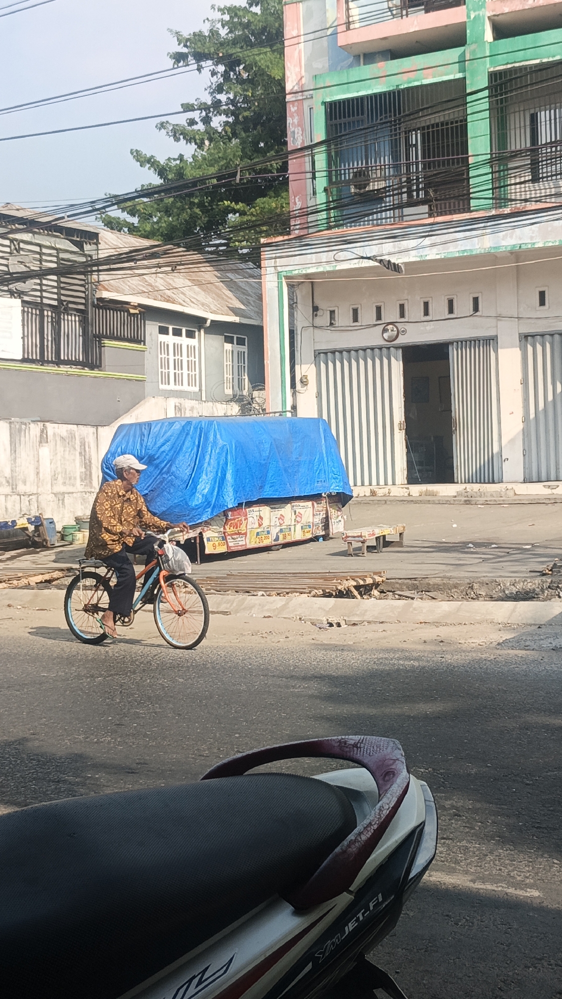 An elderly man with a white cap and a patterned brown shirt is riding a bicycle on a paved street in Banjarmasin, Indonesia. The bicycle has an orange frame, blue rims, and a basket on the handlebars. In the background, there is a building with a green and pink facade and roll-up doors. A large blue tarp covers a display of goods, which appear to be packaged items with some text visible. Power lines crisscross above the street and buildings. The scene suggests a typical urban street environment in the city.