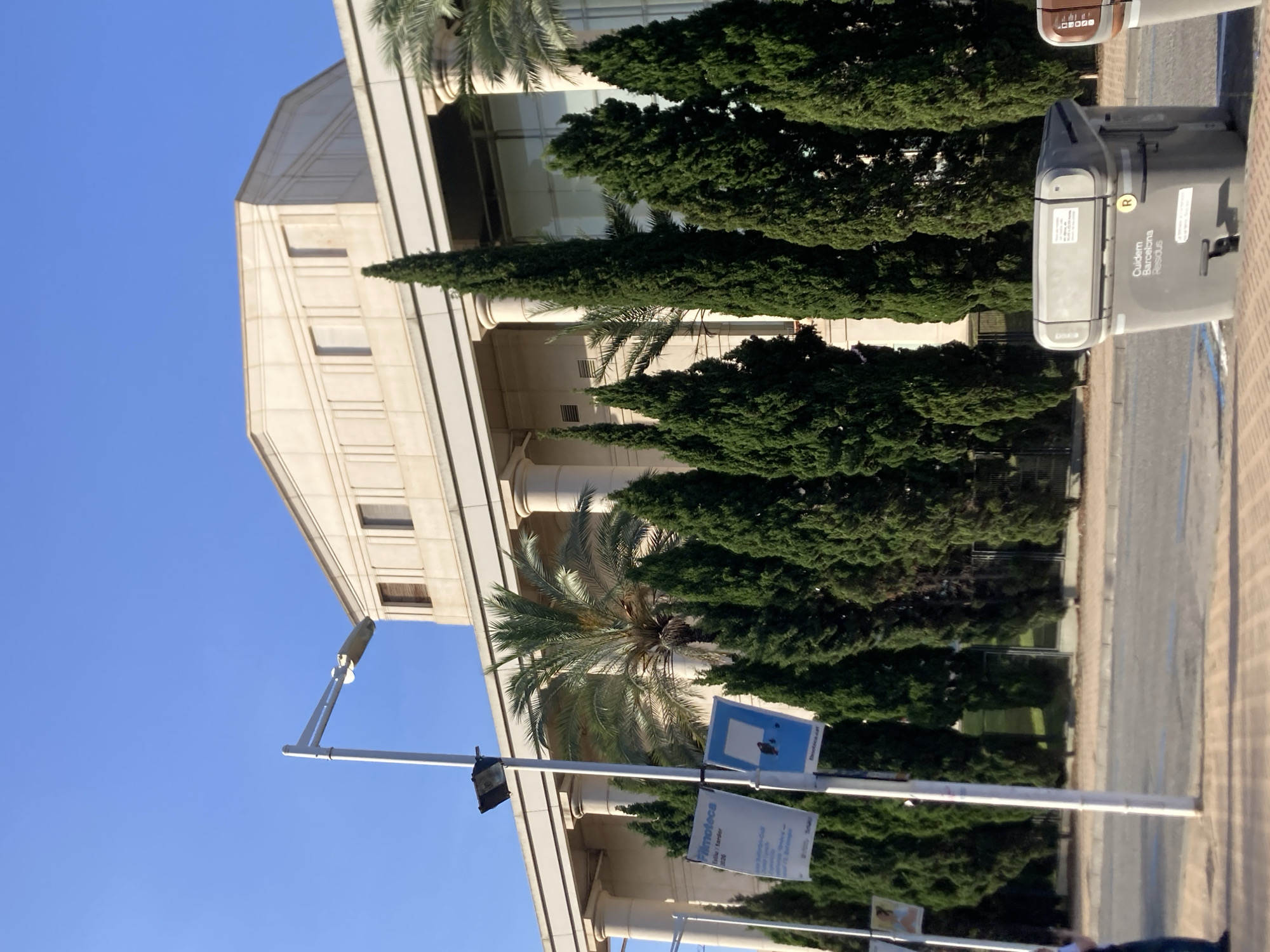 This media file captures an outdoor scene with a building, trees, and a street. The building appears to be modern with a light-colored facade, featuring prominent columns and windows. A line of manicured green trees, including cypress-like trees and a palm tree, stands in front of the building.
In the foreground, there's a street with a paved sidewalk. A street light with a modern design is visible, extending over the sidewalk. A gray public waste bin with text indicating "Guidem Barcelona Residus" is situated on the sidewalk. There are also two blue and white signs attached to poles, with some text visible on them, one of which reads "Filmoteca".
The sky is clear and blue, suggesting a sunny day. The time of day appears to be daytime, likely mid-morning or afternoon due to the bright sunlight and clear sky. The setting is an urban environment, specifically identified as Barcelona, Spain, as indicated by the text on the waste bin and the signs. No people are clearly depicted or actively engaged in any discernible activity within the frame, though the presence of urban infrastructure suggests it's a public area. The overall impression is a well-maintained public space in a city.
