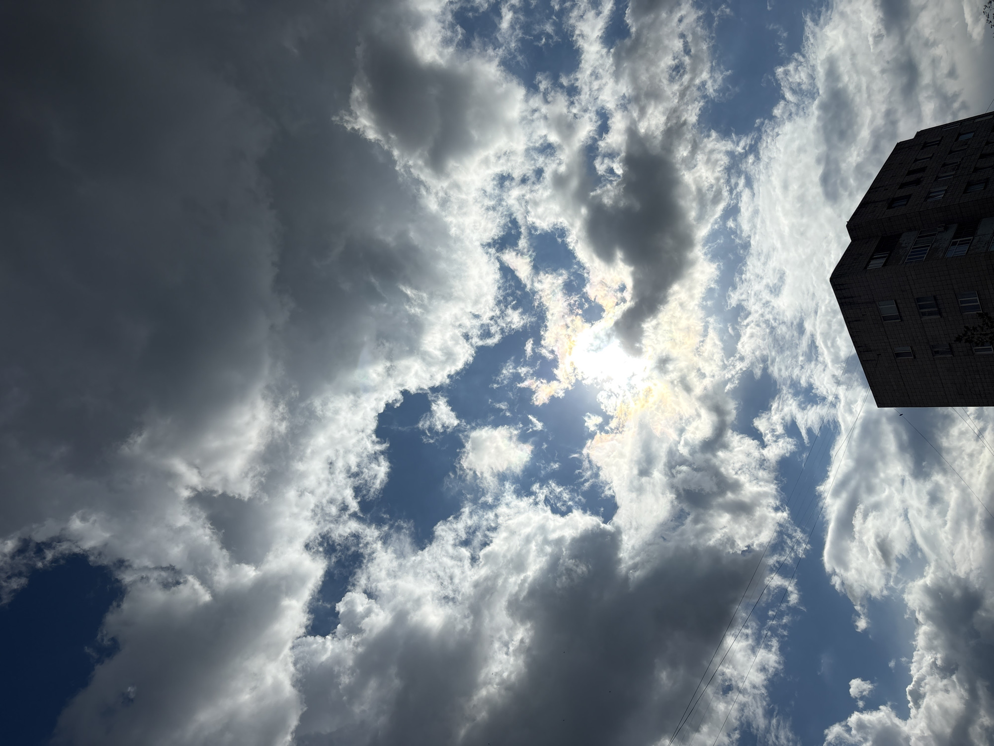 A low-angle, full shot captures a dramatic sky filled with a mix of dark, heavy clouds and bright, sunlit patches over a city building in Pavlodar, Kazakhstan. The sun is partially obscured by clouds in the upper center of the frame, casting a bright glow and creating lens flare. Electrical wires are visible stretching across the sky in the upper left and lower left quadrants. In the lower right corner, the upper section of a multi-story, concrete apartment building is visible, characterized by numerous windows. The overall atmosphere is moody, with the dynamic interplay of light and shadow in the clouds creating a sense of impending weather change.