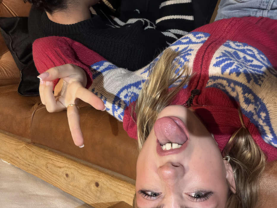 The image depicts an indoor scene in La Clusaz, France, featuring two individuals on a brown leather tufted couch. A young Caucasian female is the primary subject, positioned on her back with her head tilted to the left. She has light-colored hair and eyes, with her mouth open and tongue extended, displaying a playful expression. Her right hand (viewer's left) is raised, forming a "V" sign with extended index and middle fingers. A thin gold-colored ring is visible on her right ring finger, and her fingernails have white tips. She wears a multi-colored knit sweater or cardigan predominantly red, white, and blue, featuring geometric patterns and a visible zipper. Light-colored denim or similar fabric is partially visible below her waist. To the right and behind the female, the upper torso and a portion of the head of a second individual are visible. This person has dark, curly hair and is wearing a black knit sweater with white horizontal stripes on the lower visible section. A wooden armrest or panel is partially visible on the left side of the frame.