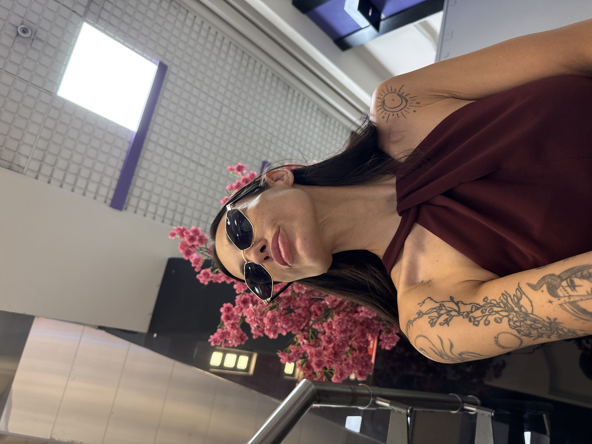 The image features an individual, likely a woman, posing indoors in what appears to be a modern commercial or public building in Pleven, Bulgaria. She has long dark hair and is wearing stylish sunglasses with dark lenses and gold frames, paired with a dark red top featuring a draped neckline. Her head is tilted, and she looks towards the camera with a confident expression, her lips slightly pouted.
Distinctive tattoos are visible on both of her arms. Her right shoulder/upper arm displays a sun and crescent moon design encircled by small stars. Her left arm is adorned with intricate floral and vine-like patterns, as well as other detailed and abstract tattoos extending down the forearm.
The background reveals architectural elements typical of a contemporary indoor space. A bright, large square light fixture illuminates a grid-patterned ceiling. Other structural components in shades of purple and dark grey/black are also visible. A prominent display of vibrant pink artificial flowers adds a decorative element. In the foreground, metallic railings suggest the presence of an escalator or staircase. A small security camera can be seen in the upper left corner, indicating a public setting. The time of day is not discernible due to the artificial lighting. There is no visible text in the image.