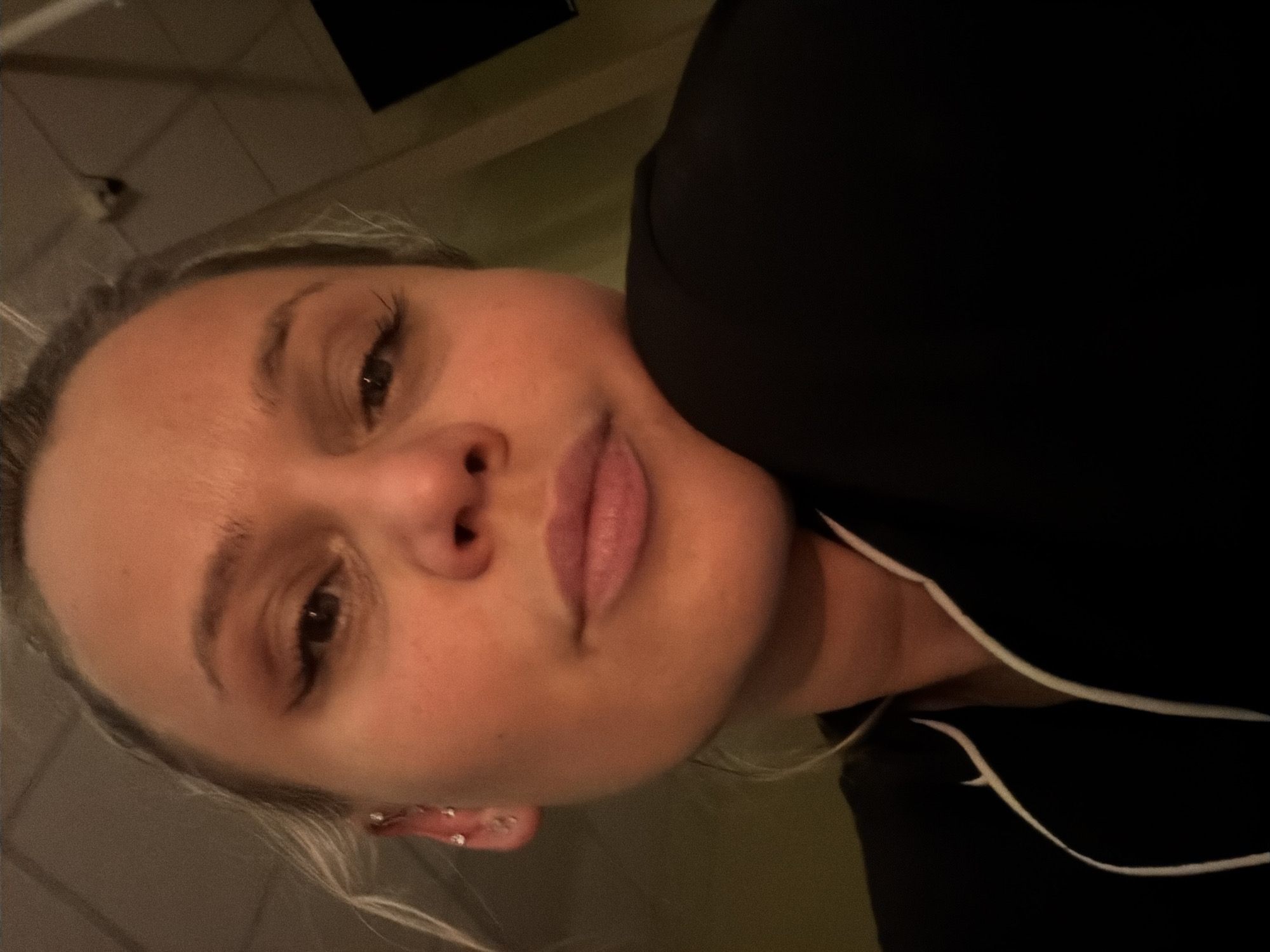 This image depicts a close-up, high-angle selfie of a single adult female subject. Her head is tilted slightly, with her right cheek resting on her right hand or arm, which is out of frame. She possesses light-colored hair, pulled back, with several small, clear stud and dangle piercings visible in her left ear. Her eyes are open, with visible eyeliner application on her upper eyelids, and she is looking directly at the camera with a neutral to slightly pensive expression. Her lips are a muted pink shade. She is wearing a dark-colored garment, possibly a top or pajamas, featuring a prominent white piping detail around the collar or lapel.
The environment is an indoor setting with artificial illumination. The background on the left side of the frame features a muted olive-green wall. The ceiling, visible in the upper portion, appears to have a grid or panel design, typical of suspended ceiling tiles, and a white, possibly recessed, light fixture is visible. Towards the upper right, a dark, rectangular object, consistent with a television screen or monitor, is partially visible against a darker wall or section. No specific landmarks or unique identifiers are visible within the image to confirm a precise location within the city of Sarapul, Russia.