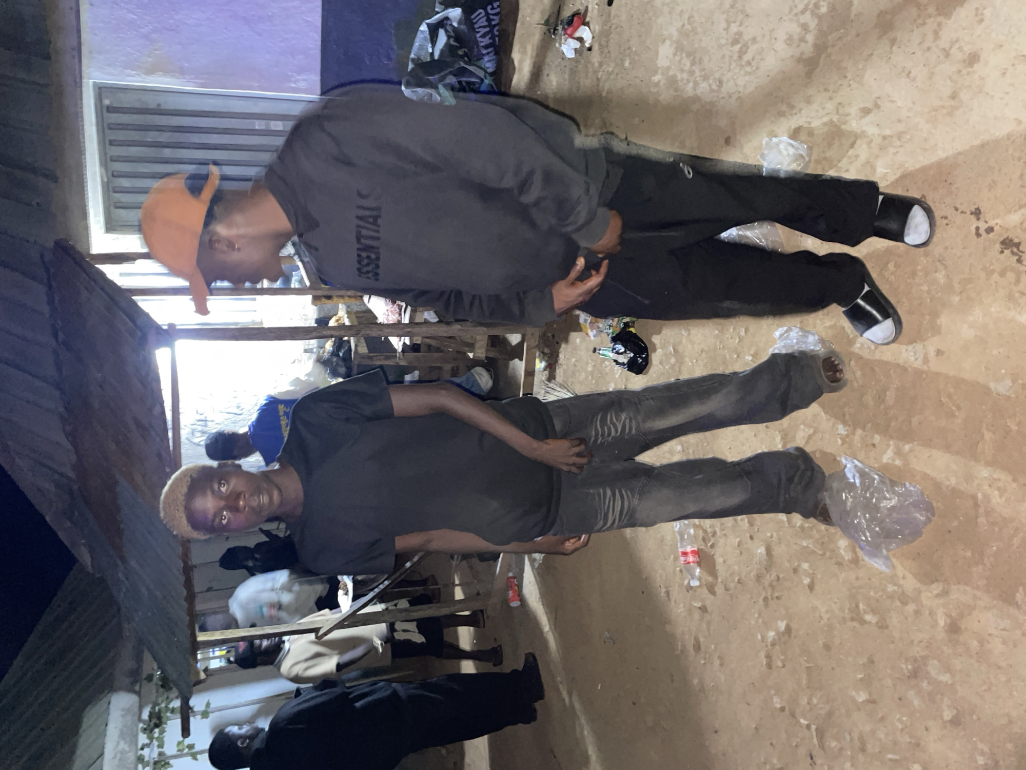 The image shows two men in an outdoor setting that could be in Durimi, Nigeria. The man in the foreground wears a black t-shirt and dark gray jeans. He has blonde hair and appears to be looking at the camera. The other man is wearing an orange baseball cap, dark grey hoodie, and black pants. Both men have plastic bags placed around their feet. There is trash and a bottle on the ground.