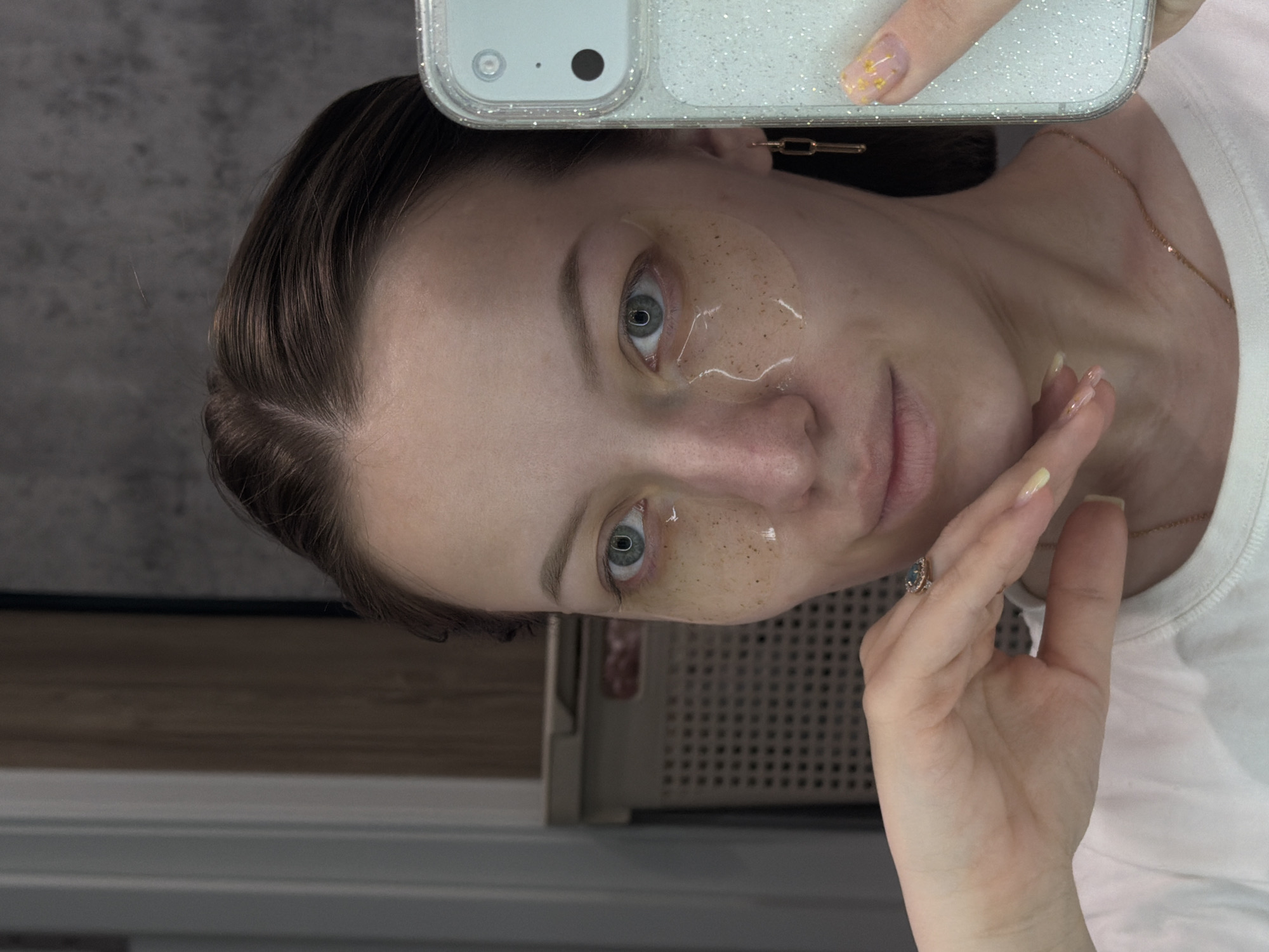 A young woman is captured in a mirror selfie, holding a smartphone above her head, its glittery white case reflecting light. The image is oriented vertically, showing the woman's head and upper torso. Her dark brown hair is styled away from her face, revealing her forehead. She has light blue-green eyes and is wearing translucent, light brown under-eye patches, which appear to contain small dark specks, suggesting a skincare routine.
She has a neutral to slightly pensive expression, looking directly into the camera's reflection. A delicate gold chain necklace is visible around her neck, resting on a white t-shirt. Her right hand, adorned with a ring featuring a light blue or grey gemstone, is positioned near her chin, with neatly manicured nails painted in a pale yellow or gold shade, possibly with glitter or gold leaf accents. A gold chain-link earring is visible on her right ear.
The background suggests an indoor setting. To the upper left, a dark, textured wall or surface is visible. To the right, behind her shoulder, is a light-colored structure with a grid pattern, possibly a ventilation unit, radiator cover, or part of a household appliance. Below her head, a wooden surface can be seen. No visible text is present in the image. The location is Reshetnikovo, Russia.