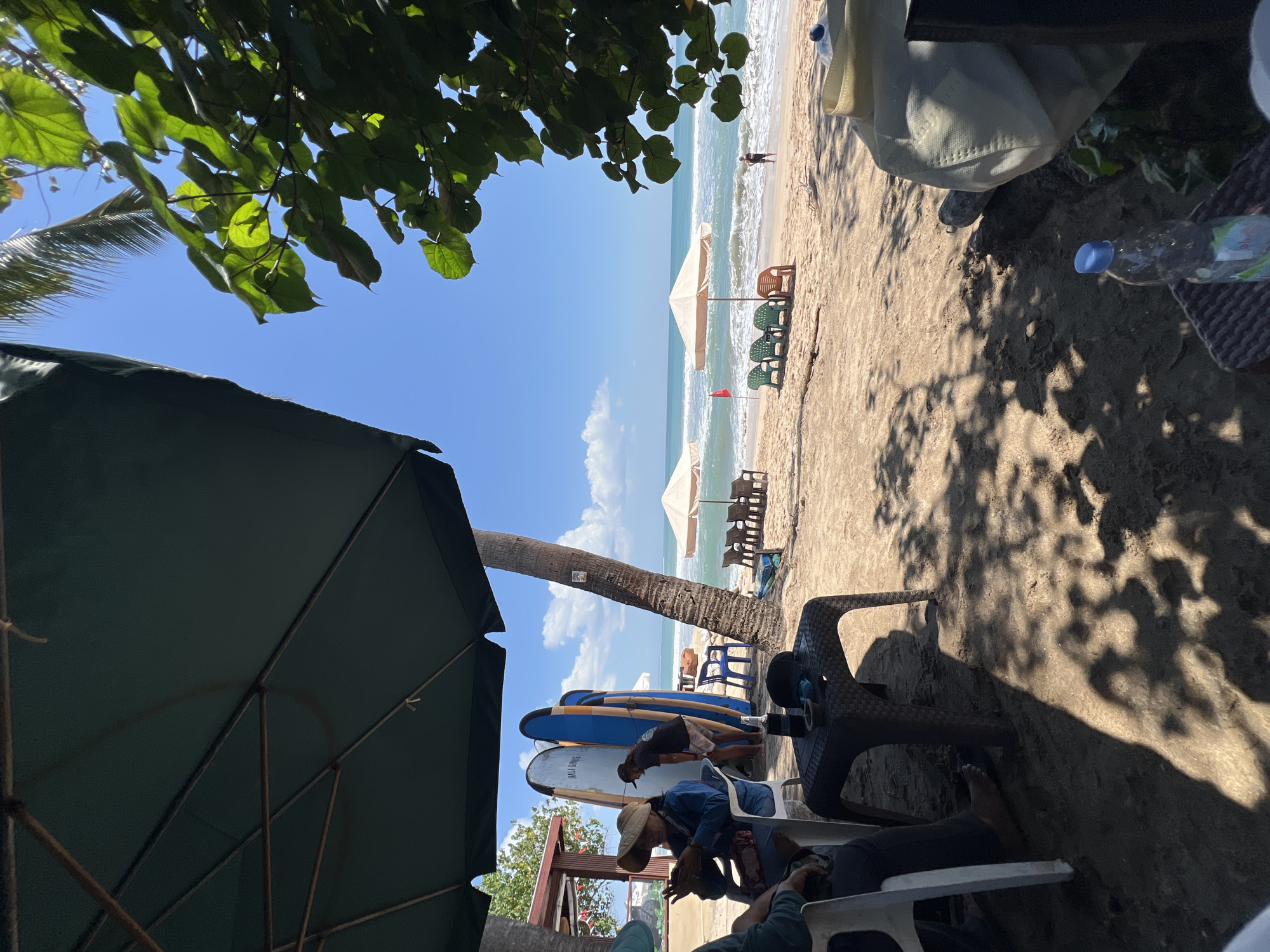 This image depicts a beach scene in Kuta, Indonesia, on a sunny day with a clear blue sky and scattered clouds. In the foreground, two people are seated at tables under a large green umbrella, with their legs and parts of their bodies visible. One person is wearing a straw hat and appears to be looking at their phone. A dark plastic table sits between them, with a bottle of water and other items on it.
Behind the seated individuals, several surfboards are leaning against a palm tree. Further back, a man in a black t-shirt and shorts is interacting with the surfboards. The sandy beach stretches towards the ocean, where gentle waves are rolling in. Several empty beach chairs and umbrellas are set up on the sand. In the distance, a lone figure is standing by the water's edge. Lush green foliage is visible in the upper right corner, framing the scene. The overall atmosphere is relaxed and indicative of a beachside establishment.