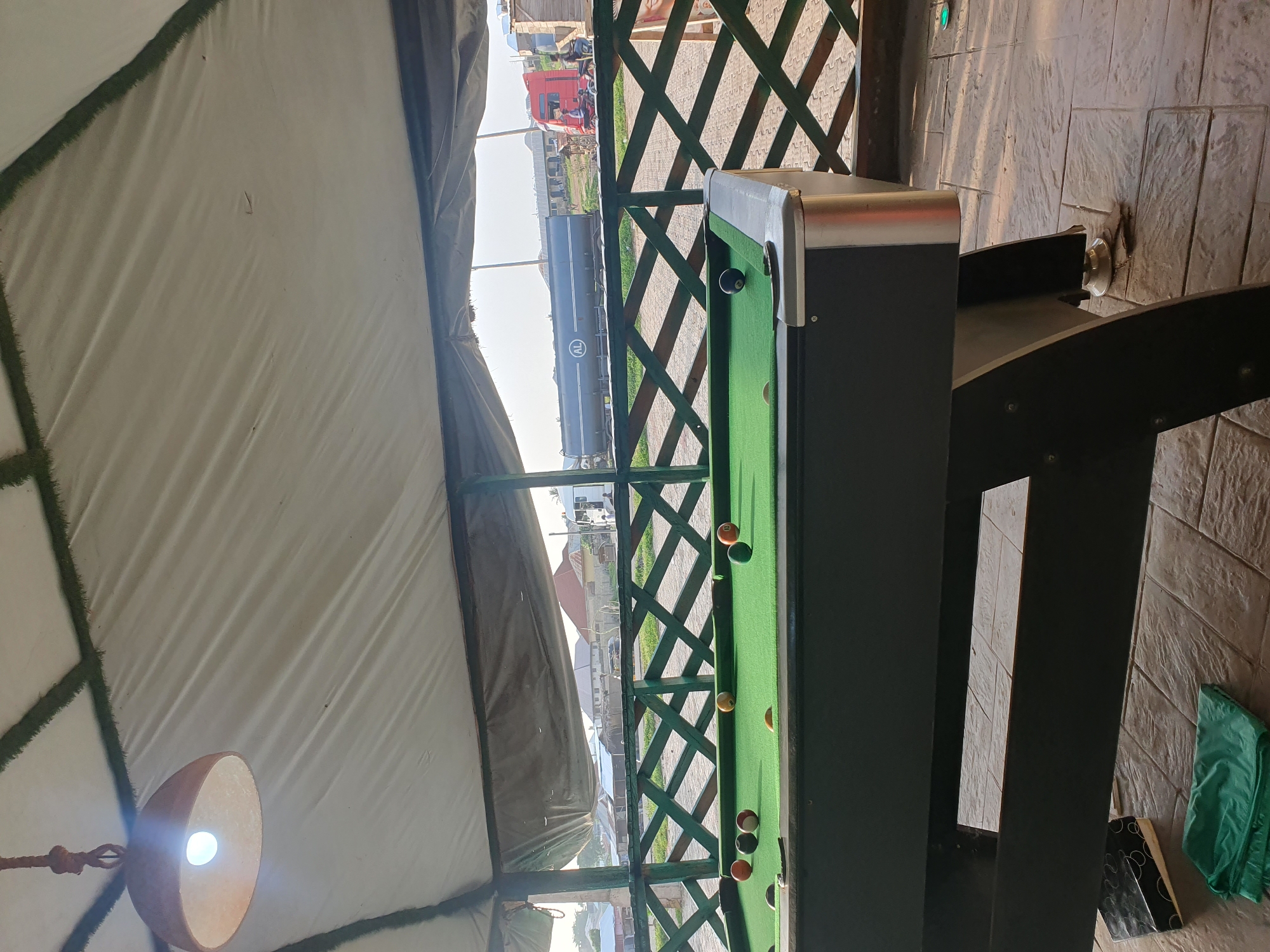 An outdoor covered area, possibly a bar or relaxation spot in Lokoja, Nigeria, features a green pool table with several billiard balls scattered across it. The table is situated on a tiled floor, and in the background, a fence with a lattice design separates the area from a road. Beyond the fence, several buildings and a large tanker truck are visible, indicating an urban or semi-urban setting. The sky is bright, suggesting daytime. A hanging lamp with a light bulb is suspended from the covered ceiling.