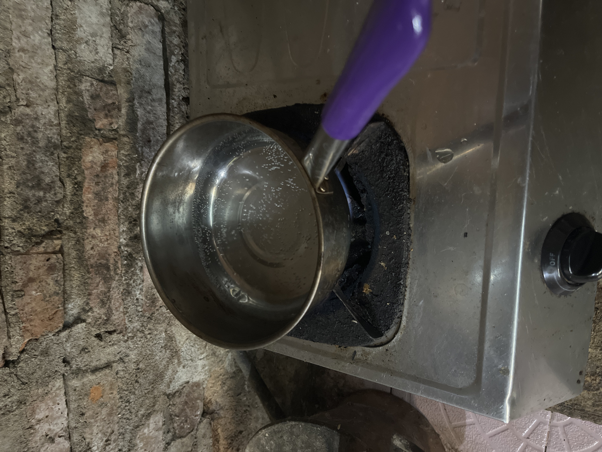 A stainless steel pot filled with bubbling water sits on a gas stove, its purple handle extending towards the viewer. The stove is situated in front of a rough, exposed brick wall. To the left of the stove, a dark, conical object is partially visible. The scene appears to be in a rustic kitchen, possibly in a residential area of Bumi Mulyo, Indonesia, indicated by the presence of a simple gas stove and brick wall construction common in some Indonesian households. The lighting suggests an indoor setting, possibly in the evening or a dimly lit room.