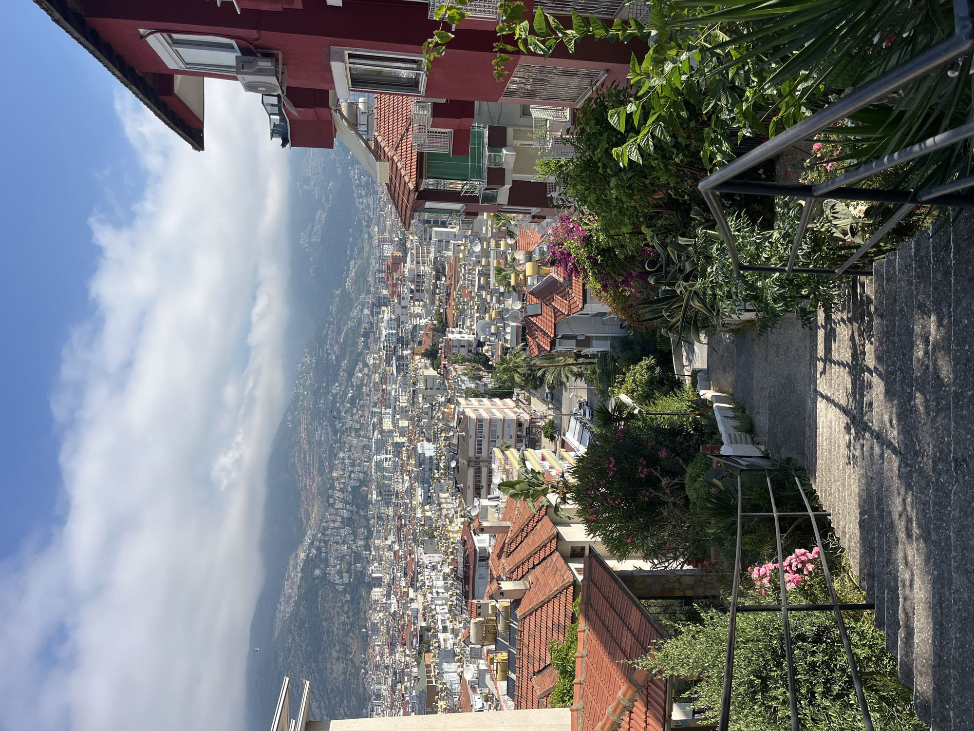 This panoramic image captures a vast cityscape cascading down a mountainside under a bright, partly cloudy sky. The scene is viewed from a higher vantage point, looking over numerous buildings in what appears to be Alanya, Turkey.
In the foreground, on the left, parts of two multi-story buildings with red facades and red-tiled roofs are visible. One building features windows and an external air conditioning unit. On the right, a stone staircase with a dark metal railing descends, framed by lush green vegetation. This greenery includes bushes, palm leaves, and flowering plants with pink and purple blossoms, adding vibrant color to the foreground. Sunlight casts clear shadows across the steps and railing, indicating it is daytime, likely mid-morning or afternoon.
The midground and background are dominated by a dense urban sprawl of residential and commercial buildings, many featuring distinctive red-tiled roofs and white or light-colored facades. These structures extend far into the distance, climbing the slopes of large, verdant mountains that form the backdrop. Some clouds hang low over the mountain range, creating a soft, misty appearance. The sky above is a clear blue with scattered white clouds. The overall impression is one of a vibrant coastal city nestled between mountains and the sea, though the sea itself is not visible in this particular shot.
There is no discernible human activity or visible text in the image.