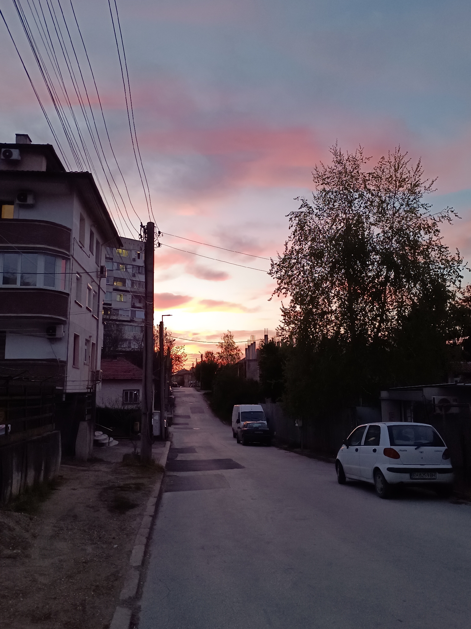 This media file presents a serene residential street scene in Pleven, Bulgaria, captured during either sunrise or sunset. The sky is a prominent feature, displaying a beautiful blend of soft pinks, oranges, and light blues from the setting or rising sun, with scattered clouds.
On the left, several multi-story residential buildings line the street. The closest building features balconies and external air conditioning units, with lights visible in some windows. Numerous power lines crisscross the upper left part of the image, connecting to a utility pole that also supports a streetlamp. Further down the street on the left, another taller residential building is visible. The ground immediately adjacent to the buildings consists of a dirt and patchy grass area.
The asphalt street, which also incorporates sections of concrete paving, leads into the distance. On the right side of the street, a large, full-leaved tree stands silhouetted against the vibrant sky, dominating the mid-ground. Further down, more trees and bushes are visible.
Several vehicles are parked along the street. On the right, a white hatchback, identified by its design as likely a Daewoo Matiz, is parked with its rear facing the viewer. Its license plate, "EH 6259 BH", is clearly visible. Further down the street on the right, a darker van is parked. Another white van is parked on the left side of the street, further into the background.
The scene conveys a calm and peaceful ambiance, typical of a quiet residential area during the twilight hours. The weather appears clear, allowing for the striking natural light display in the sky.