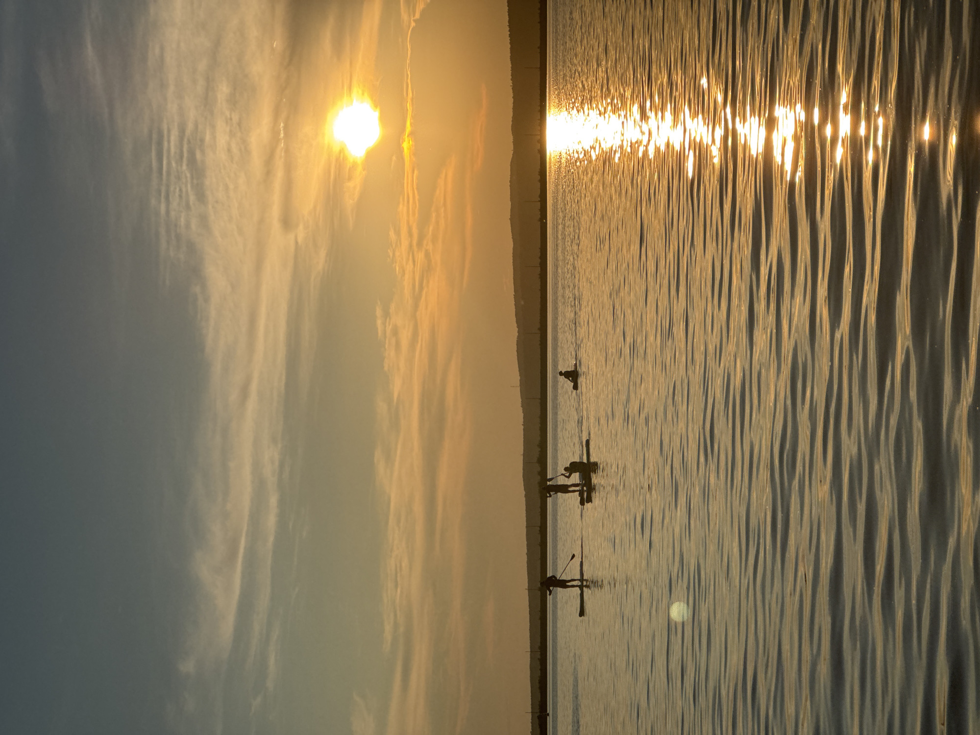 The image depicts a serene sunset over the Kama River in Chaikovsky, Russia. Silhouettes of four individuals on paddleboards are visible on the water, enjoying the twilight. The sun, a bright orb, is setting in the upper right portion of the frame, casting a golden glow across the sky and reflecting a shimmering path on the rippling water. The background features a distant, hazy shoreline with rolling hills and a few power lines. The sky is a gradient of soft blues and oranges, with wispy clouds catching the last rays of sunlight. The overall atmosphere is tranquil and picturesque, capturing a moment of peaceful recreation at dusk.