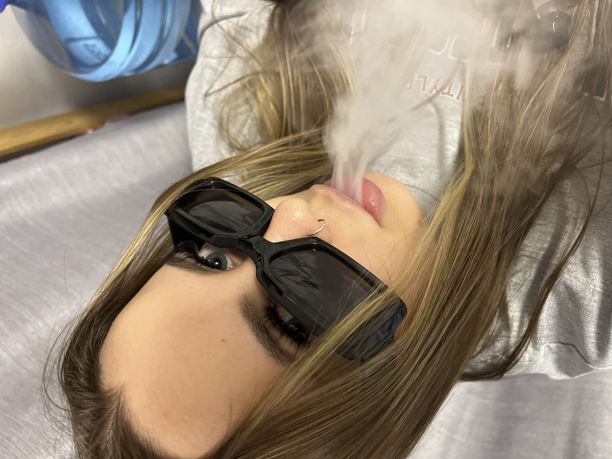 A young woman lies on her back, wearing sunglasses, with smoke emanating from her mouth. She has light brown hair and a nose ring. She is wearing a grey sweatshirt. The background includes a blue object and a wooden rod. The location is not discernible from the provided image.