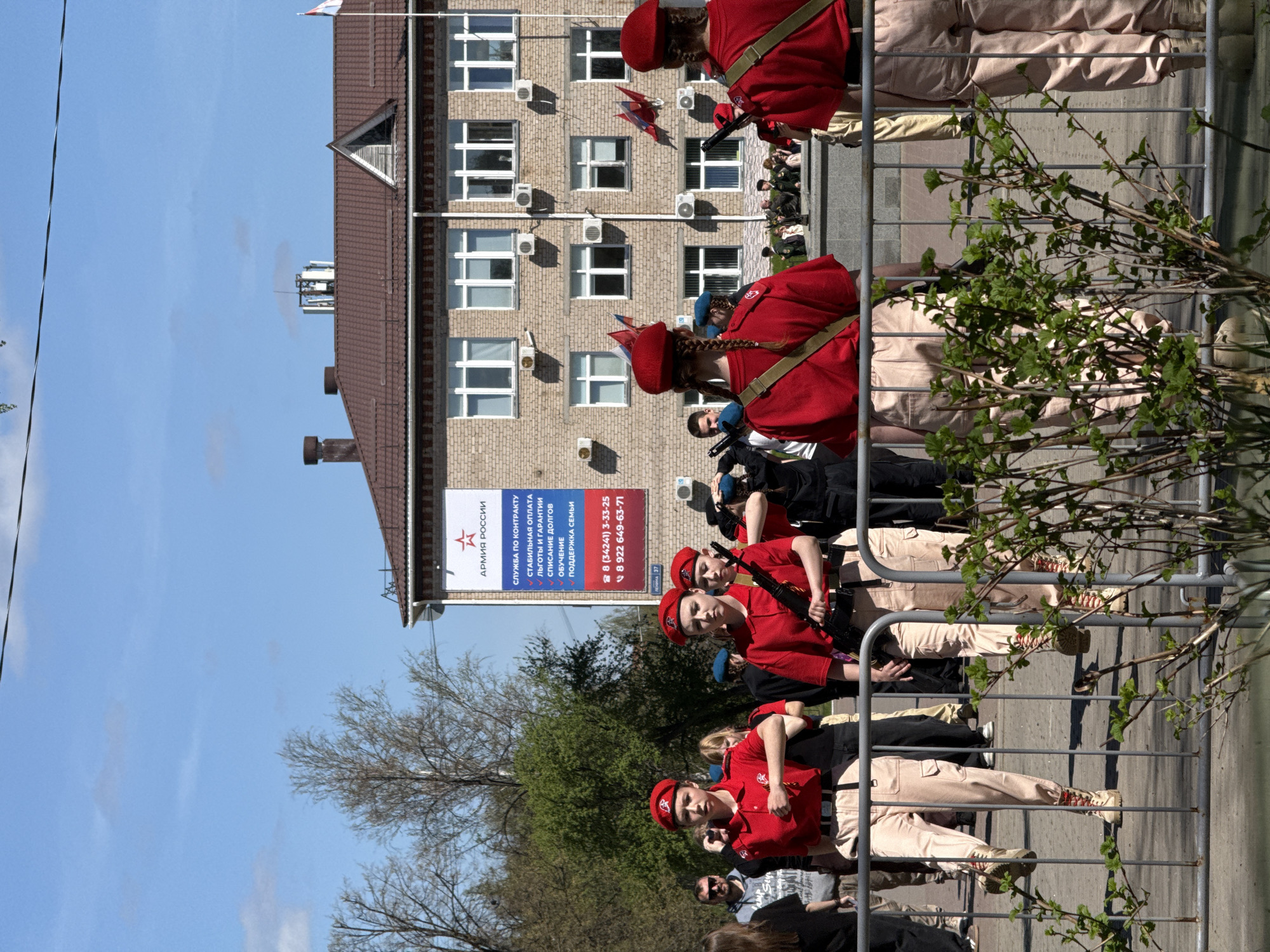 The image shows a group of children dressed in military uniforms with red berets standing behind a metal fence. They are holding what appear to be toy rifles. Behind them is a multi-story brick building with a brown roof. A large banner on the building displays text in Russian, including phone numbers: 8 (34241) 3-33-25 and 8 922 649-63-71, as well as information about serving in the Russian army. According to the sign "Shkola 37", the location is likely a school in the city of Chaikovsky, Russia. The sky is blue and there are trees in the foreground.