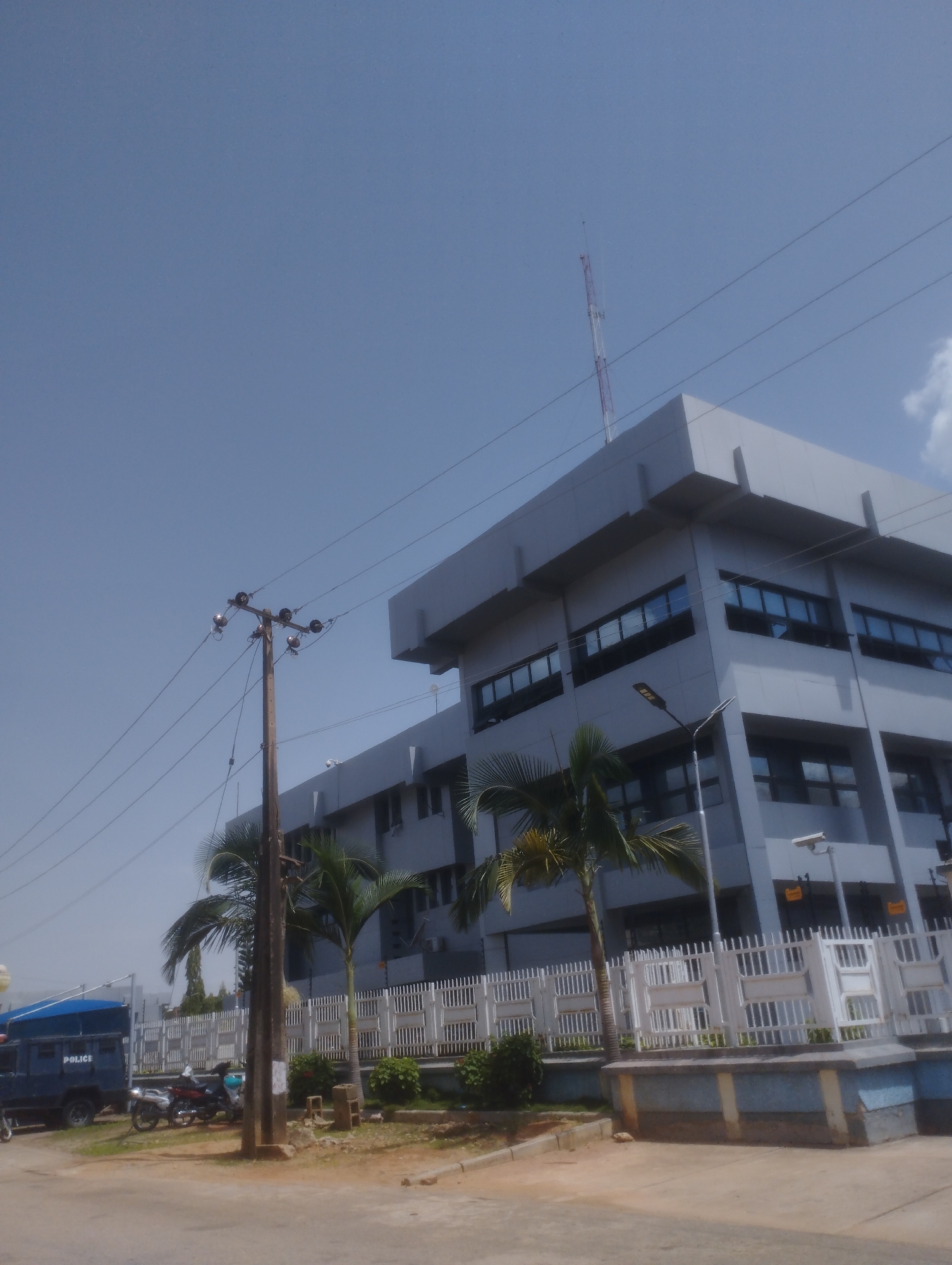 The image depicts an outdoor scene in what appears to be an urban or semi-urban environment in Jos, Nigeria. The primary subject is a modern, multi-story building with a light gray facade and dark tinted windows. The building has a sharp, angular design. In front of the building, there is a white picket fence with decorative elements. Several palm trees are visible, suggesting a warm climate.
To the left of the building, a utility pole with electrical wires and insulators stands prominently. A dark blue police van with "POLICE" written on its side is parked behind the utility pole. Two motorcycles are parked nearby, and a person is visible on one of them.
In the background, a tall communication tower or antenna is visible against a clear blue sky. The lighting suggests it is daytime, likely mid-morning or afternoon, with bright sunlight casting shadows. The weather appears to be clear and sunny. The presence of the police vehicle and the building's architecture might suggest a government or security-related facility. The overall atmosphere is neutral and functional.