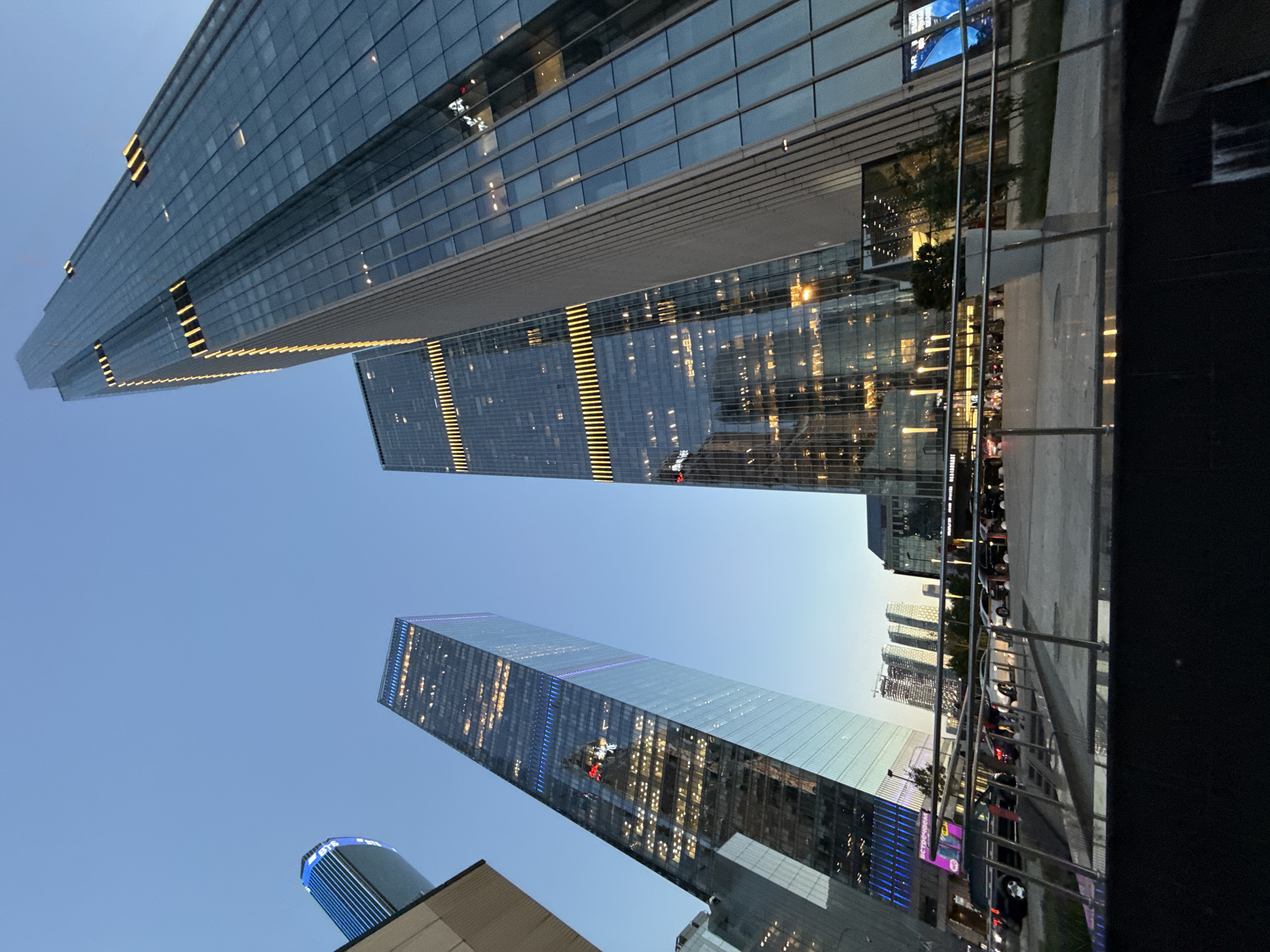 This photograph, set in Khimki, Russia, captures a low-angle perspective of a modern urban landscape during twilight, showcasing a cluster of three prominent glass-clad skyscrapers. On the right, the tallest structure is a slender, rectangular tower featuring numerous horizontal bands of illuminated yellow lights across its blue glass facade. To its left, a slightly shorter but equally modern skyscraper exhibits a glass exterior with varied light patterns, including vertical blue and purple strips along its edges and a discernible red MTS logo (a stylized 'M' within a red square with a star) on a lower, illuminated section. Partially visible on the far left is a third skyscraper characterized by a curved top and vertical blue strip lighting along its visible edge.
In the mid-ground, a paved pedestrian area extends across the scene, leading towards the base of the buildings. Several vehicles, including cars and potential taxis, are parked or in motion on an adjacent roadway. The immediate foreground features a dark, reflective surface, possibly water or a polished floor, beneath a horizontal metal railing that traverses the image. Small, illuminated streetlights are present at ground level. A white planter containing greenery is situated near the center of the pedestrian area. Various signs, including one with partially legible Cyrillic text ("встречаем") and another indicating "RESTAURANT," are visible on the lower sections of the buildings and surrounding structures, alongside a large blue display screen on the far right. The sky above transitions from a light blue at the horizon to a deeper hue overhead, with artificial building and ground lights clearly illuminated.